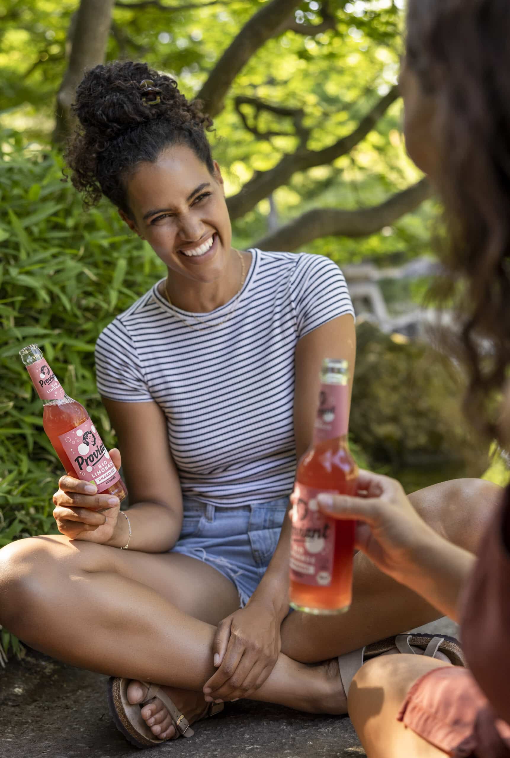 Zwei Frauen sitzen im Freien und halten Flaschen mit Limonade in der Hand. Die Frau links lächelt und trägt ein gestreiftes Hemd und Jeansshorts. Sie sitzt im Schneidersitz auf dem Boden. Die Kulisse scheint ein Park mit Grünflächen und Bäumen im Hintergrund zu sein. © Fotografie Tomas Rodriguez