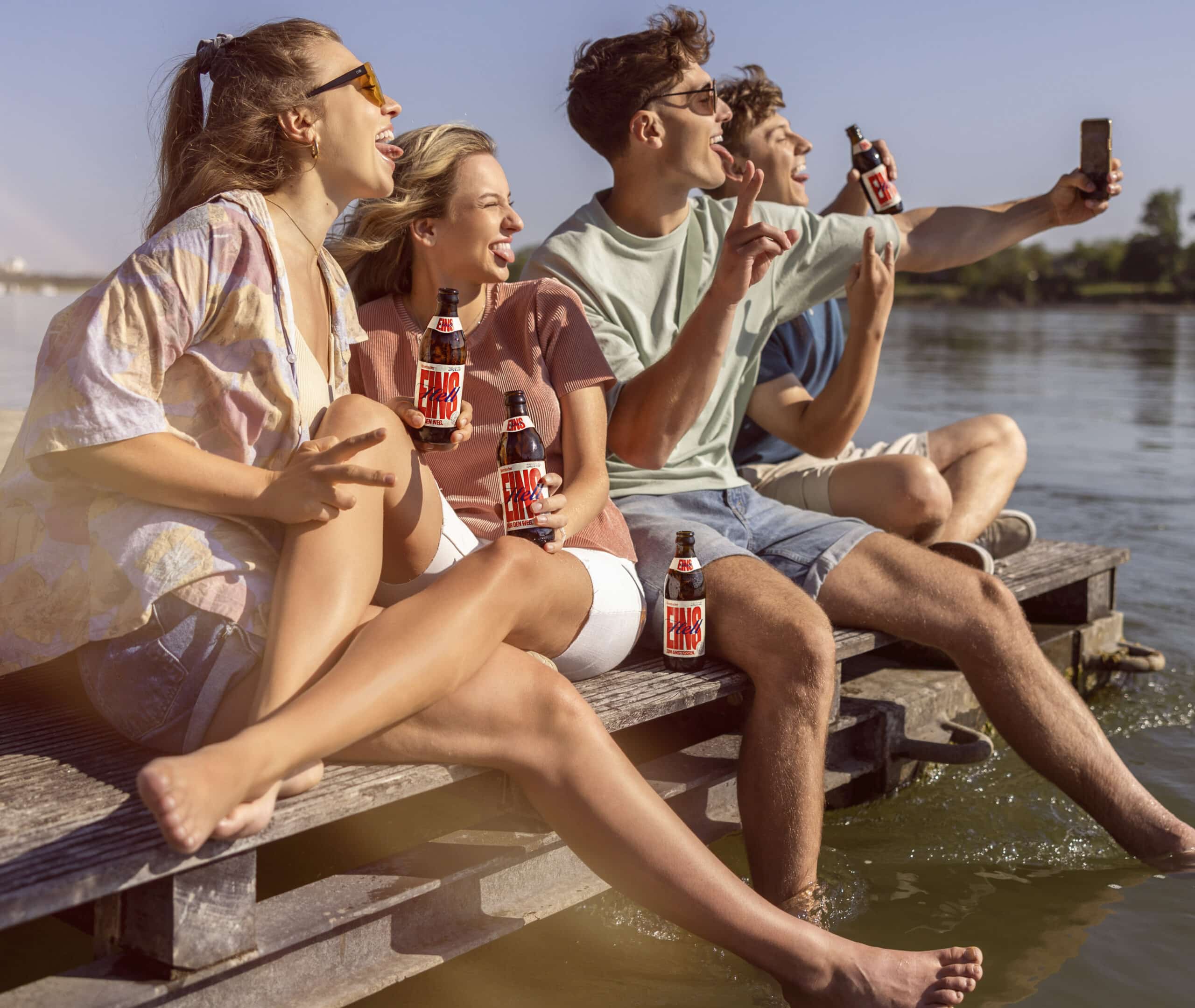 Vier junge Erwachsene sitzen am Ufer eines Sees, halten Getränkeflaschen in der Hand und lächeln. Sie sind barfuß, zwei tauchen ihre Füße ins Wasser. Eine Person macht ein Selfie. Die Szene ist sonnig und entspannt und lässt auf einen lustigen Ausflug am Wasser schließen. © Fotografie Tomas Rodriguez
