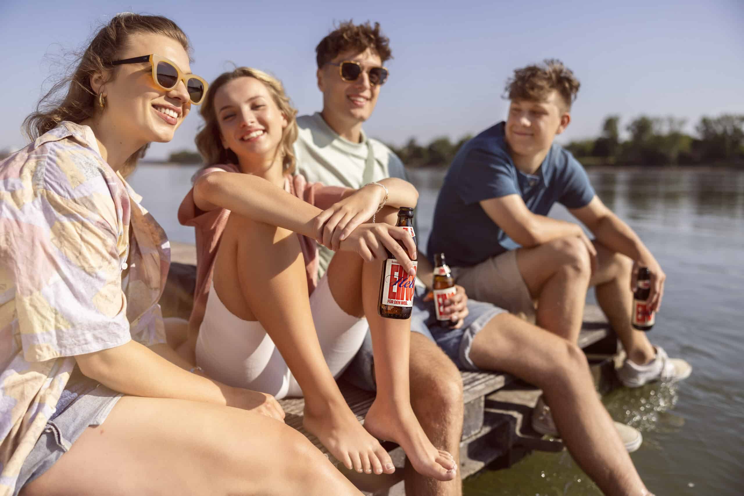 Vier Freunde sitzen an einem sonnigen Tag auf einem Holzsteg an einem See. Drei von ihnen tragen Sonnenbrillen, die Gruppe lächelt und hält Bierflaschen in den Händen. Das Wasser ist ruhig, im Hintergrund sind Bäume zu sehen. Die Atmosphäre ist entspannt und fröhlich. © Fotografie Tomas Rodriguez