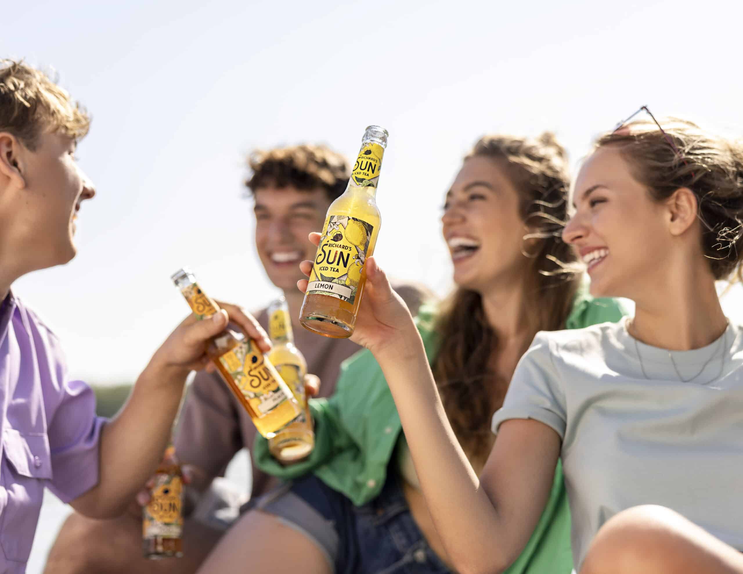 Eine Gruppe von vier Freunden mit glücklichen Gesichtern genießt Flaschengetränke mit der Aufschrift „SONNE“. Sie sind draußen bei strahlendem Sonnenschein. Die Gruppe unterhält sich fröhlich und stößt mit ihren Flaschen an. Eine Person trägt eine Sonnenbrille auf dem Kopf. © Fotografie Tomas Rodriguez