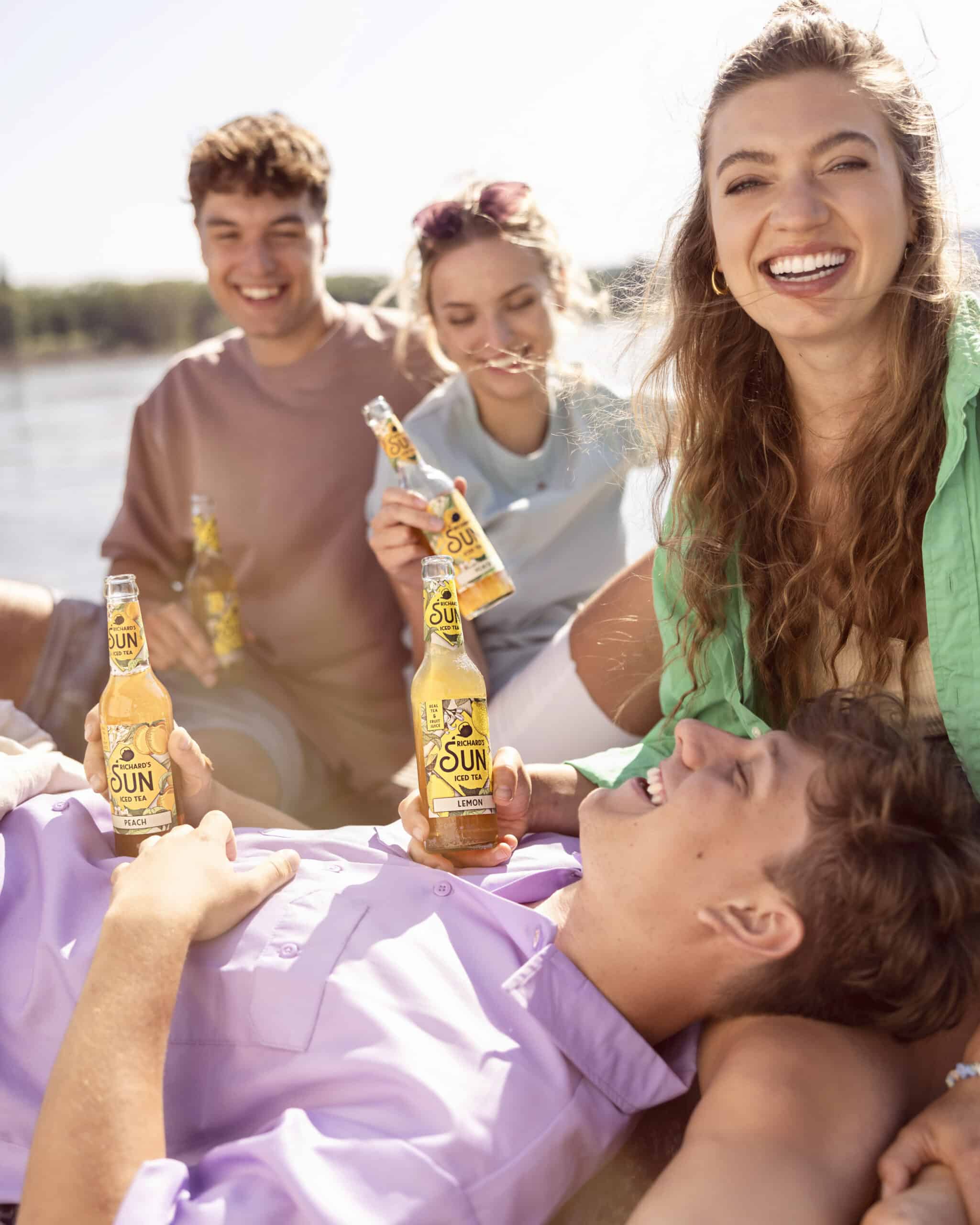Vier Menschen sitzen an einem sonnigen Tag im Freien, lächeln und genießen Getränke aus Flaschen. Eine Person liegt mit dem Kopf auf dem Schoß einer anderen Person und hält eine Flasche in der Hand. Die anderen sitzen in der Nähe und wirken alle entspannt und glücklich. © Fotografie Tomas Rodriguez