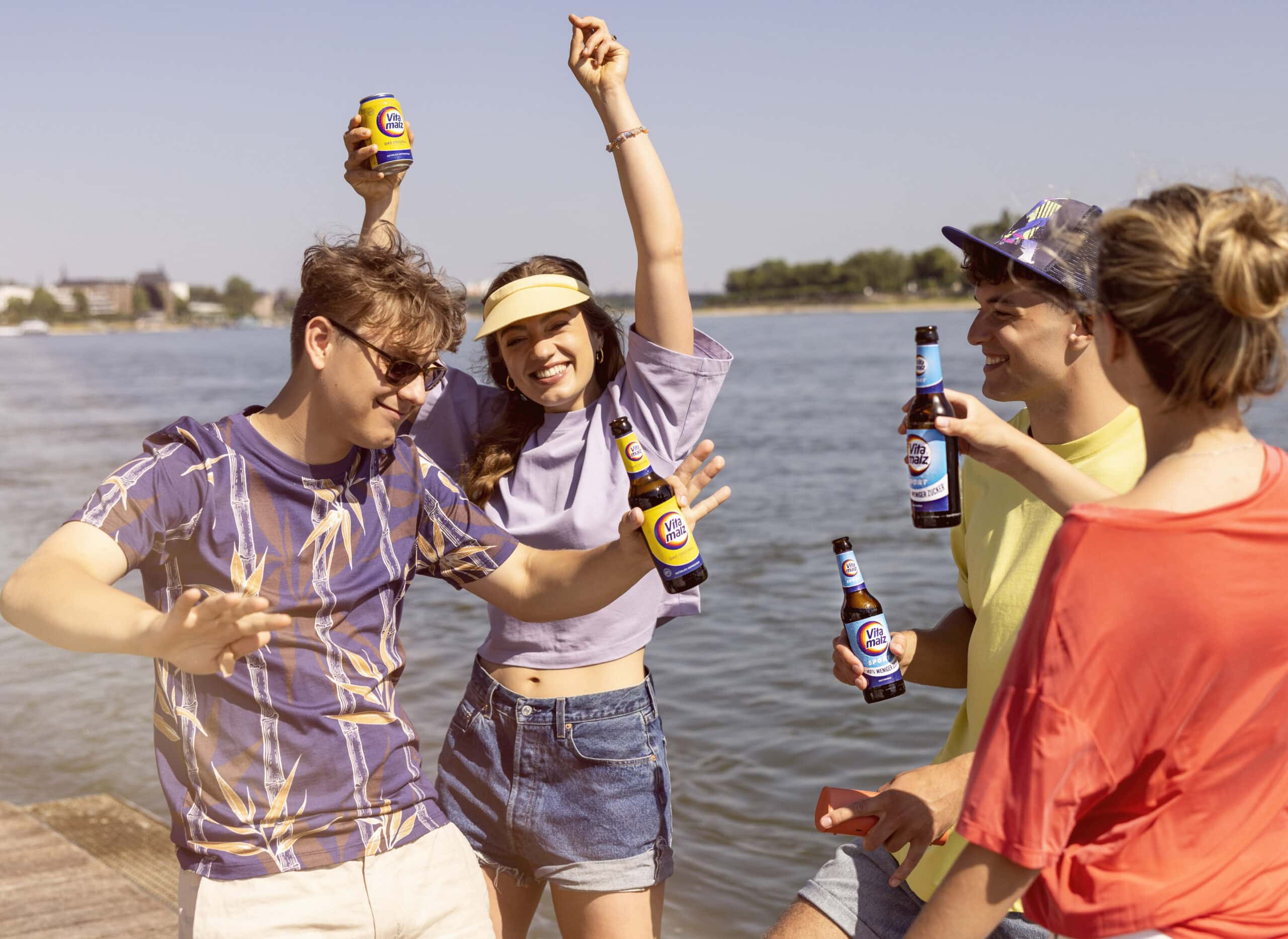 Vier Freunde genießen einen sonnigen Tag am Wasser. Eine Person im Tropenhemd tanzt, während eine andere mit Schirmmütze fröhlich ihr Getränk hebt. Eine dritte Person lacht und hält ein Bier in der Hand, und eine vierte in einem roten Hemd schaut zu. Alle halten Getränke in der Hand, im Hintergrund ein ruhiger See. © Fotografie Tomas Rodriguez