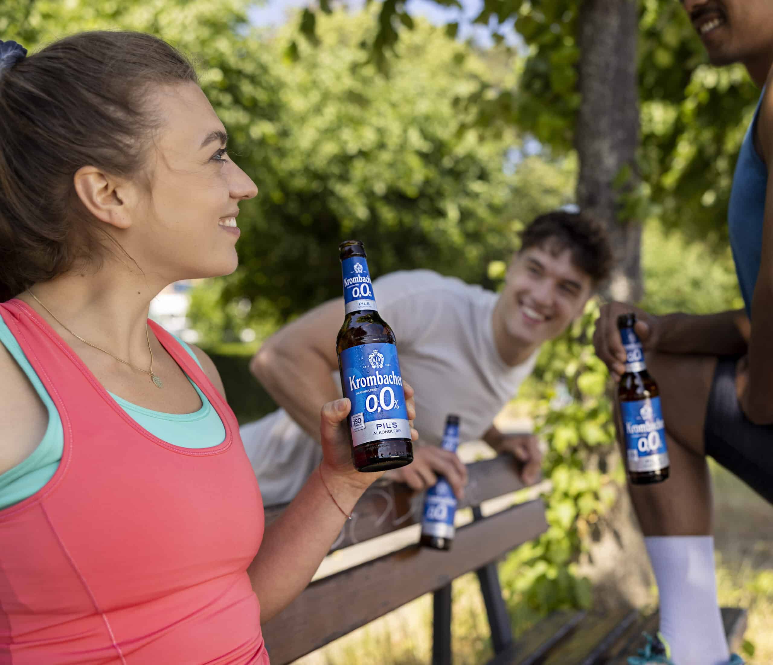 Drei Personen sitzen im Freien, lächeln und halten Flaschen Krombacher 0,0 % Pils in der Hand. Sie scheinen einen entspannten Moment zusammen auf einer Holzbank mit grünen Bäumen und einer sonnigen Kulisse zu genießen. Die Person in der Mitte ist im Fokus, während die anderen in einer entspannten Pose stehen. © Fotografie Tomas Rodriguez