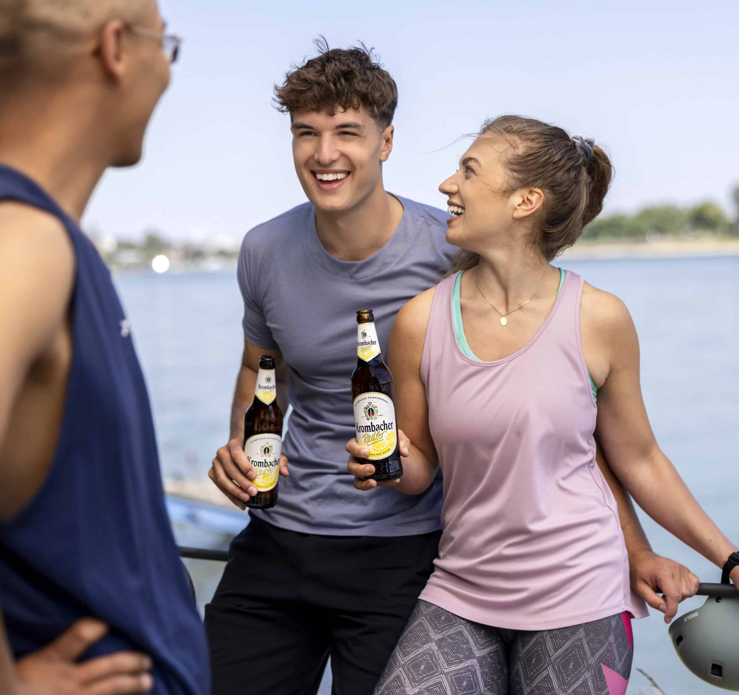 Eine Gruppe von drei Freunden lacht und plaudert am Wasser. Zwei von ihnen, ein Mann und eine Frau, halten Flaschen Krombacher Bier in den Händen. Die dritte Person, teilweise sichtbar, steht mit dem Rücken zur Kamera. Der Himmel ist klar und die Stimmung ausgelassen. © Fotografie Tomas Rodriguez