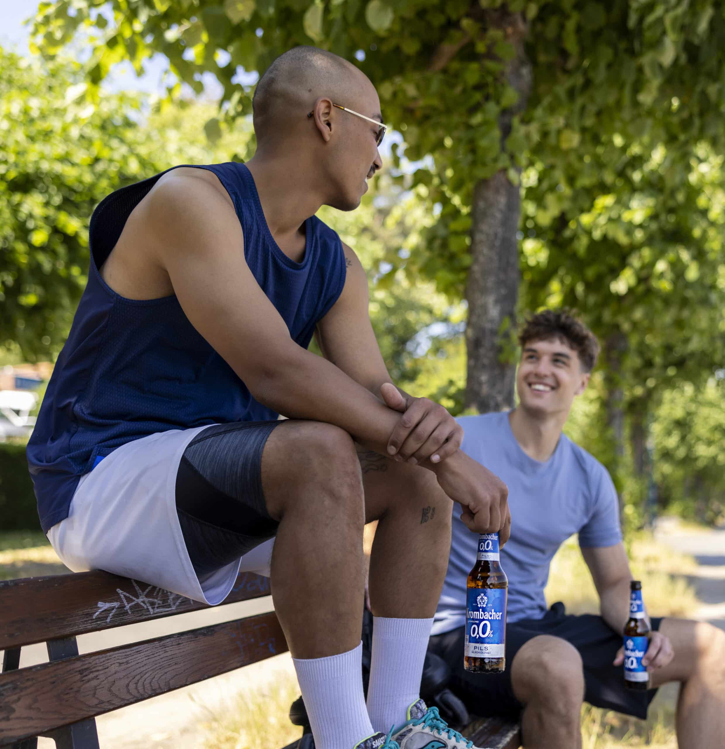 Zwei Männer sitzen an einem sonnigen Tag lachend auf einer Bank und halten Flaschen mit alkoholfreiem Bier in den Händen. Ein Mann mit rasiertem Kopf trägt eine Sonnenbrille und Sportkleidung, während der andere mit kurzen Haaren ein hellblaues T-Shirt trägt. Im Hintergrund sind Bäume und ein Weg zu sehen. © Fotografie Tomas Rodriguez