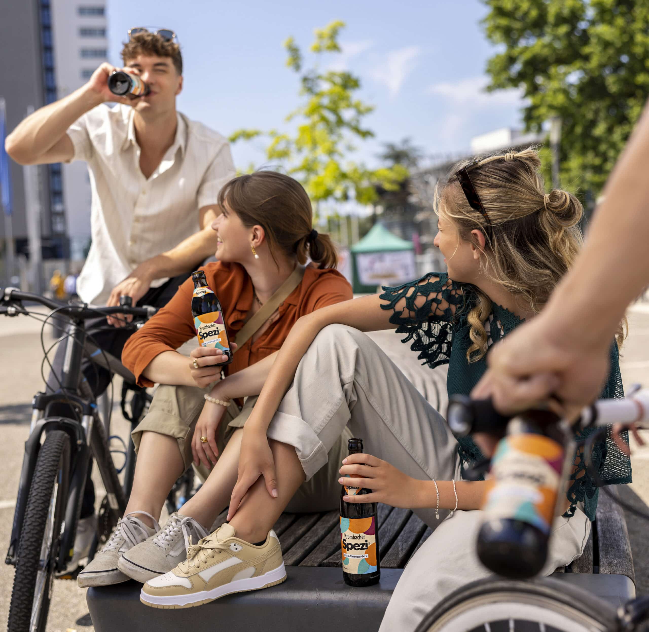 Drei junge Leute mit Fahrrädern sitzen und stehen im Freien und genießen Getränke aus Flaschen mit der Aufschrift „Spezi“. Einer nippt an einer Flasche, während die anderen ihre halten und sich in lässiger Kleidung vor einer städtischen Kulisse mit Bäumen und einem Gebäude entspannen. © Fotografie Tomas Rodriguez