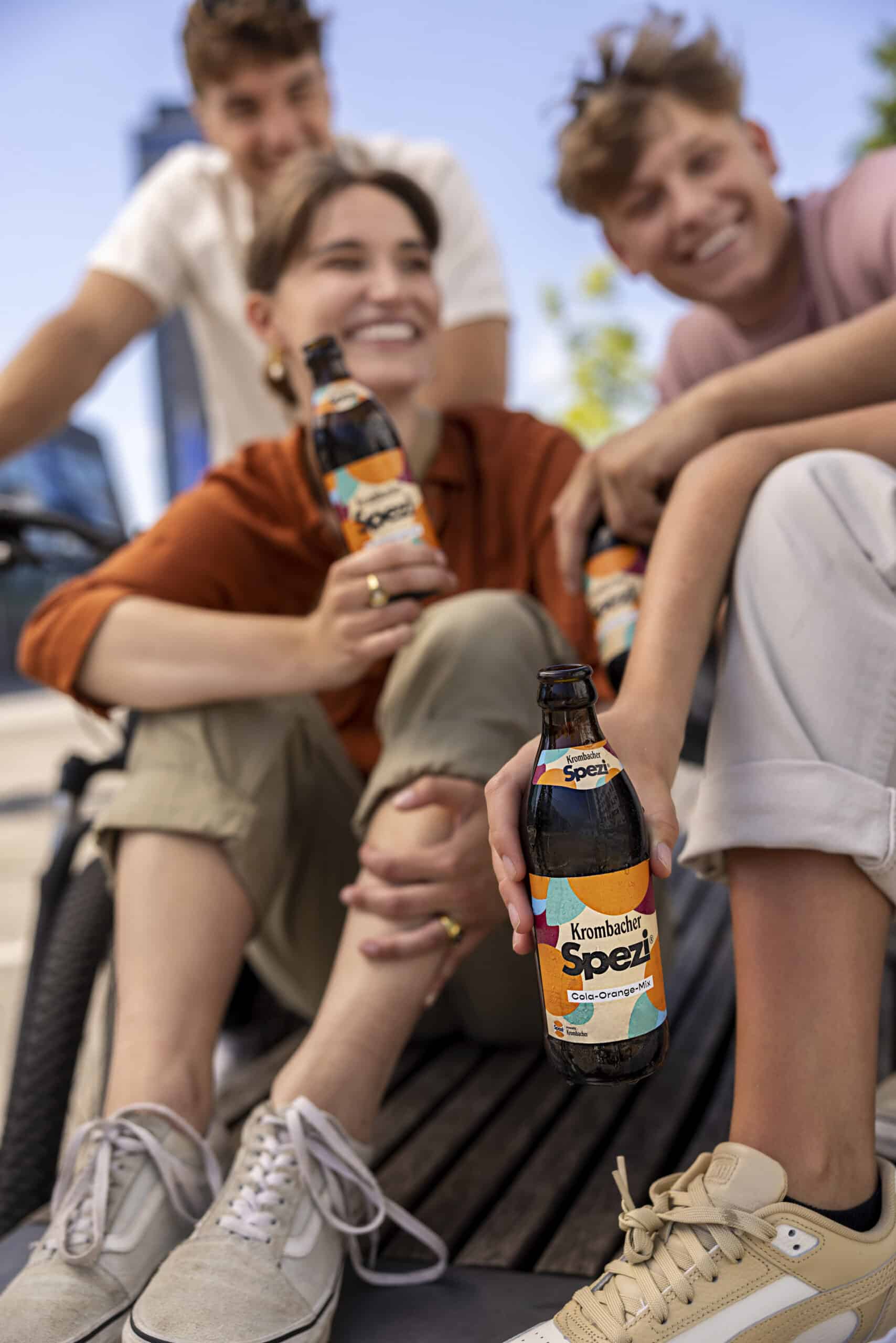Eine Gruppe von vier lächelnden Freunden genießt ihre Zeit im Freien. Jeder hält eine Flasche Krombacher Spezi in der Hand. Sie sitzen lässig, tragen legere Kleidung und wirken entspannt und glücklich. Der Hintergrund zeigt eine sonnige Stadtlandschaft. © Fotografie Tomas Rodriguez