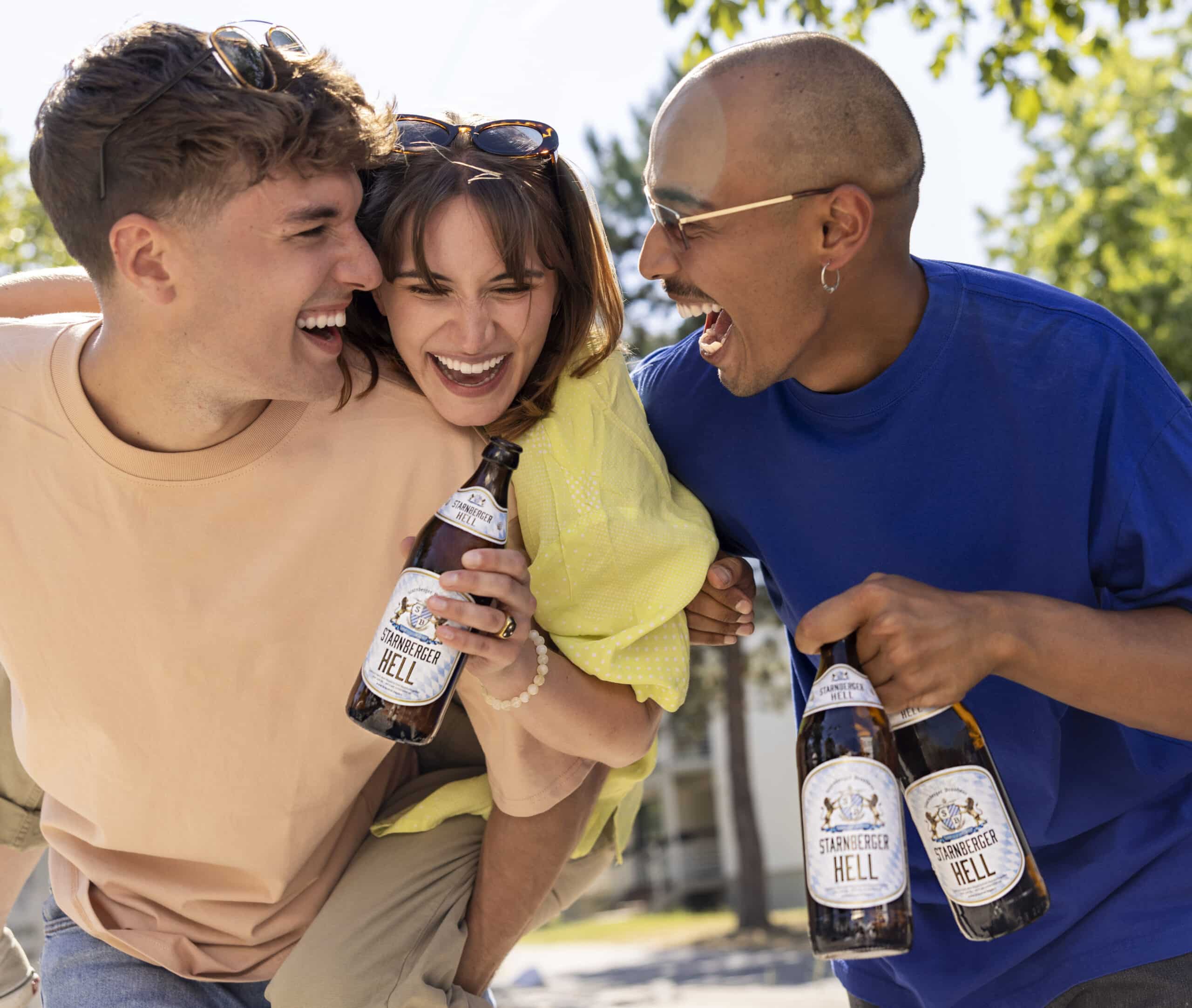 Drei Freunde verbringen einen fröhlichen Moment im Freien. Die Person links (im hellen Hemd) trägt die Person in der Mitte (im gelben Hemd), die eine Bierflasche hält, Huckepack. Die Person rechts, die eine Brille und ein blaues Hemd trägt, hält ebenfalls Bierflaschen und lacht. © Fotografie Tomas Rodriguez