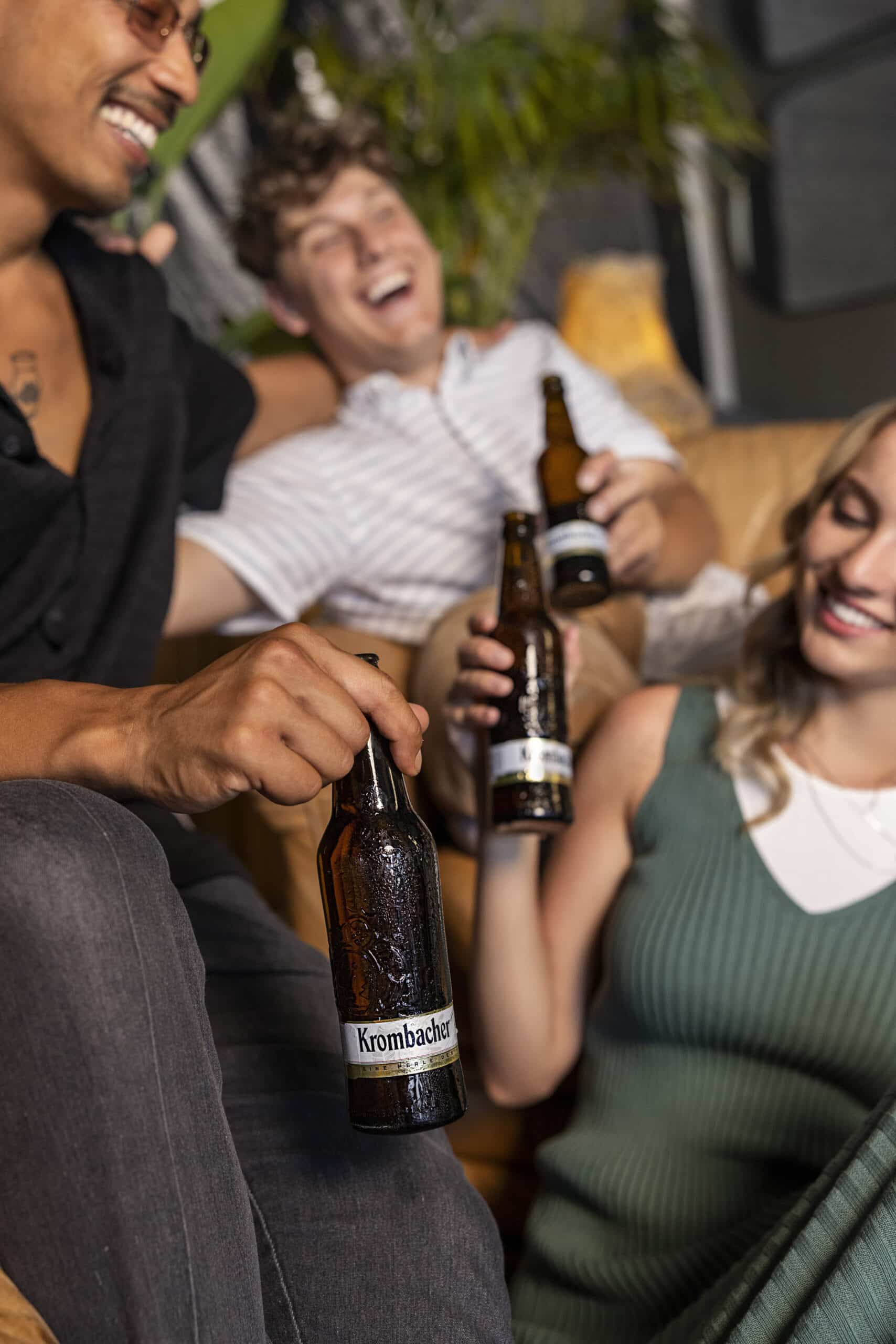 Drei Personen sitzen zusammen und lachen, während sie Flaschen Krombacher Bier in den Händen halten. Sie scheinen eine gute Zeit zu haben, wobei eine Person im Vordergrund eine Brille und ein schwarzes Hemd trägt, während die anderen beiden im Hintergrund leicht verschwommen sind. © Fotografie Tomas Rodriguez