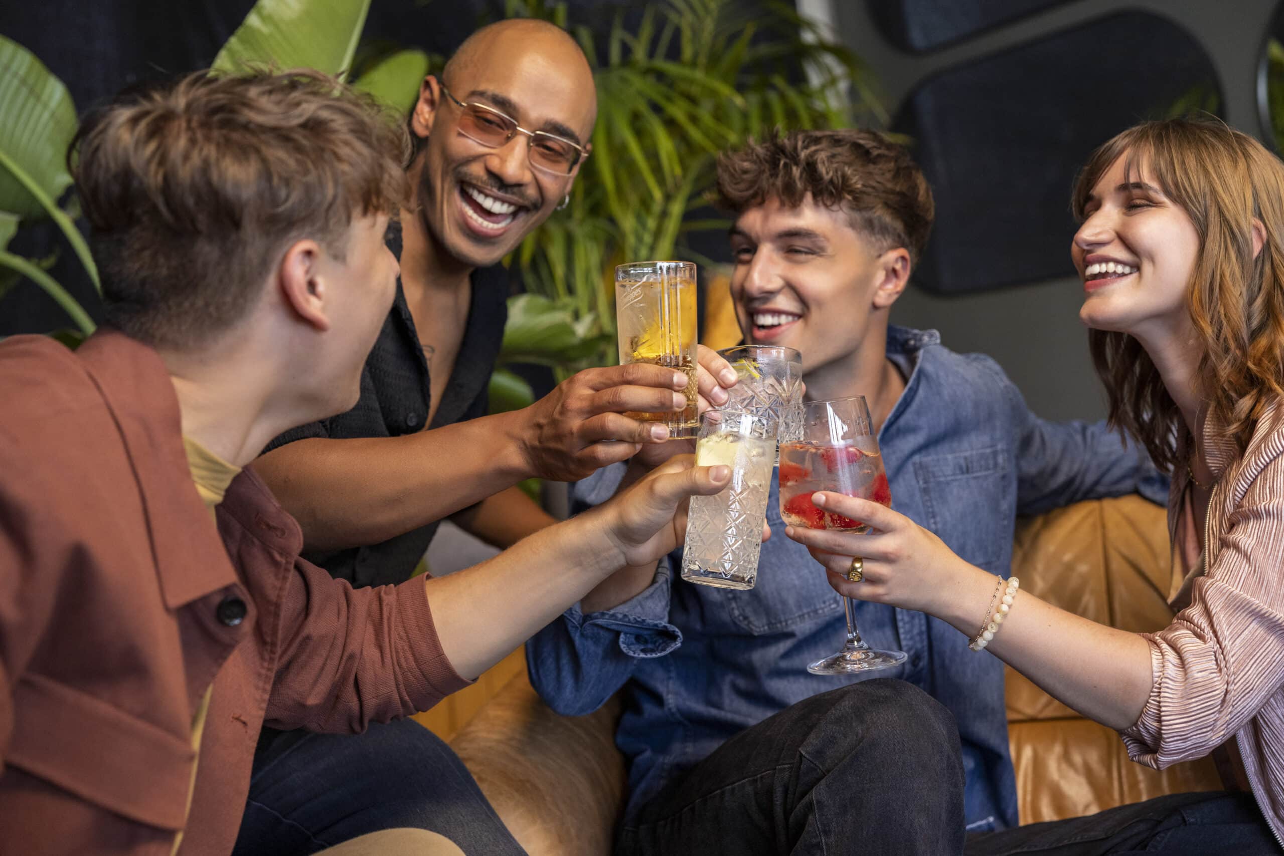 Eine Gruppe von vier Freunden, zwei Männer und zwei Frauen, sitzt auf einem Sofa, lächelt und stößt mit ihren Getränken an. Sie befinden sich in einer gemütlichen Umgebung mit grünen Pflanzen im Hintergrund. © Fotografie Tomas Rodriguez