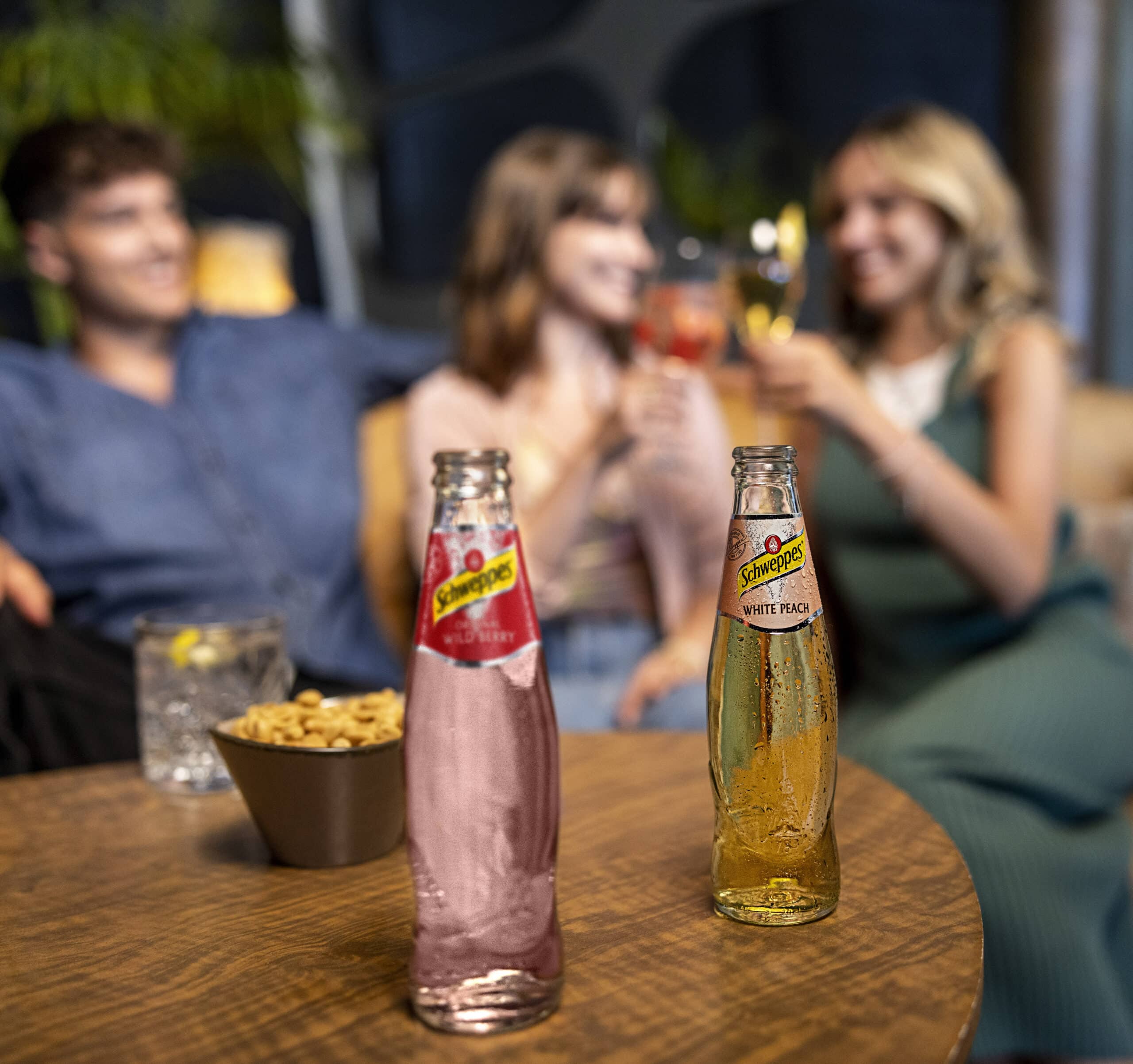 Drei Personen sitzen an einem Tisch, heben ihre Gläser und lächeln. Im Vordergrund stehen zwei Flaschen Schweppes, eine mit einer rosa Flüssigkeit mit der Aufschrift „Raspberry“ und die andere mit einer gelben Flüssigkeit mit der Aufschrift „White Peach“. Auf dem Tisch stehen außerdem eine Schale mit Nüssen und ein Getränk. © Fotografie Tomas Rodriguez