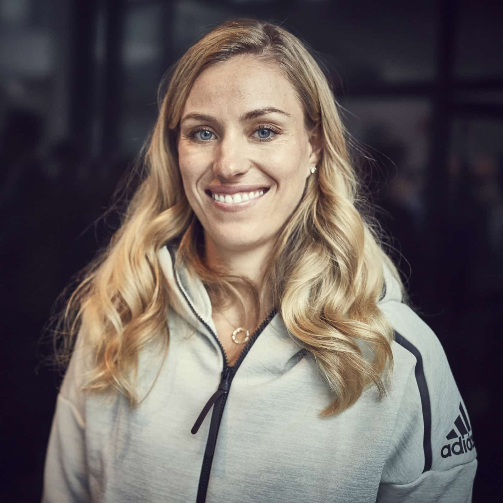 Eine Frau mit langen blonden Haaren und blauen Augen lächelt in die Kamera. Sie trägt eine hellgraue Reißverschlussjacke mit dem Adidas-Logo auf der Schulter. Der Hintergrund ist dunkel und unscharf. © Fotografie Tomas Rodriguez