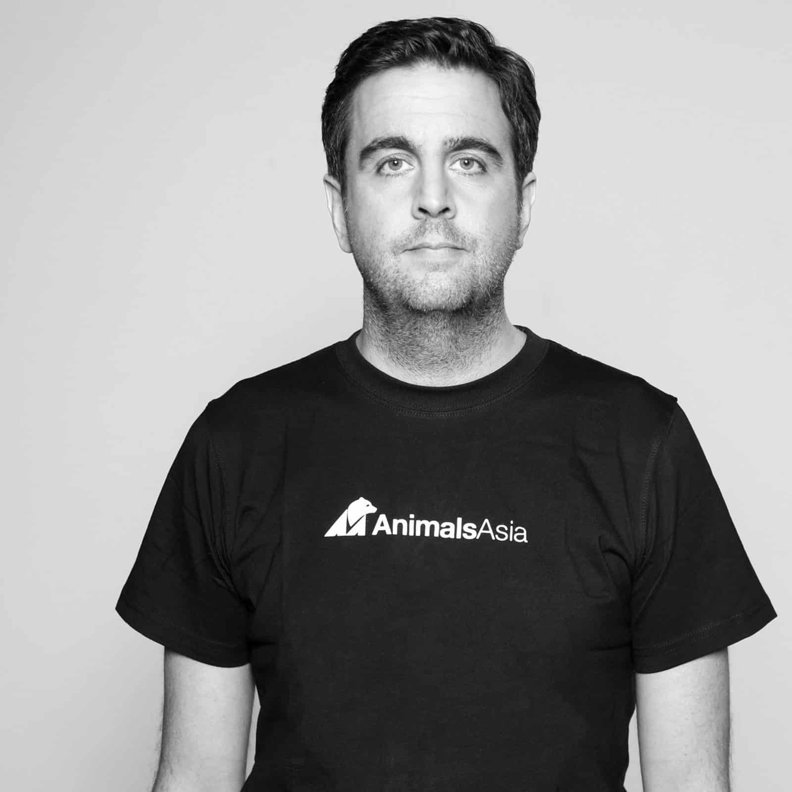 Ein Mann mit kurzen dunklen Haaren steht vor einem schlichten Hintergrund. Er trägt ein schwarzes T-Shirt mit dem Logo von „Animals Asia“. Sein Gesichtsausdruck ist neutral. © Fotografie Tomas Rodriguez