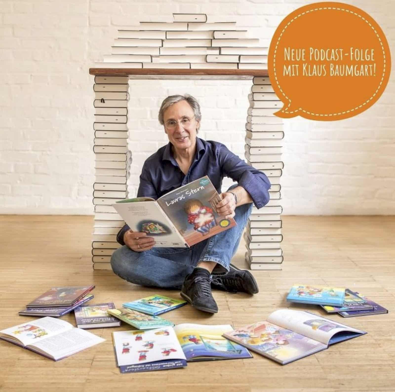 Vor einem Gerüst aus Bücherstapeln sitzt ein Mann auf dem Boden und hält ein Buch von „Lauras Stern“ in der Hand. Um ihn herum sind weitere bunte Bücher verteilt. In der oberen rechten Ecke befindet sich eine Sprechblase mit dem Text „Neue Podcast-Folge mit Klaus Baumgart“. © Fotografie Tomas Rodriguez