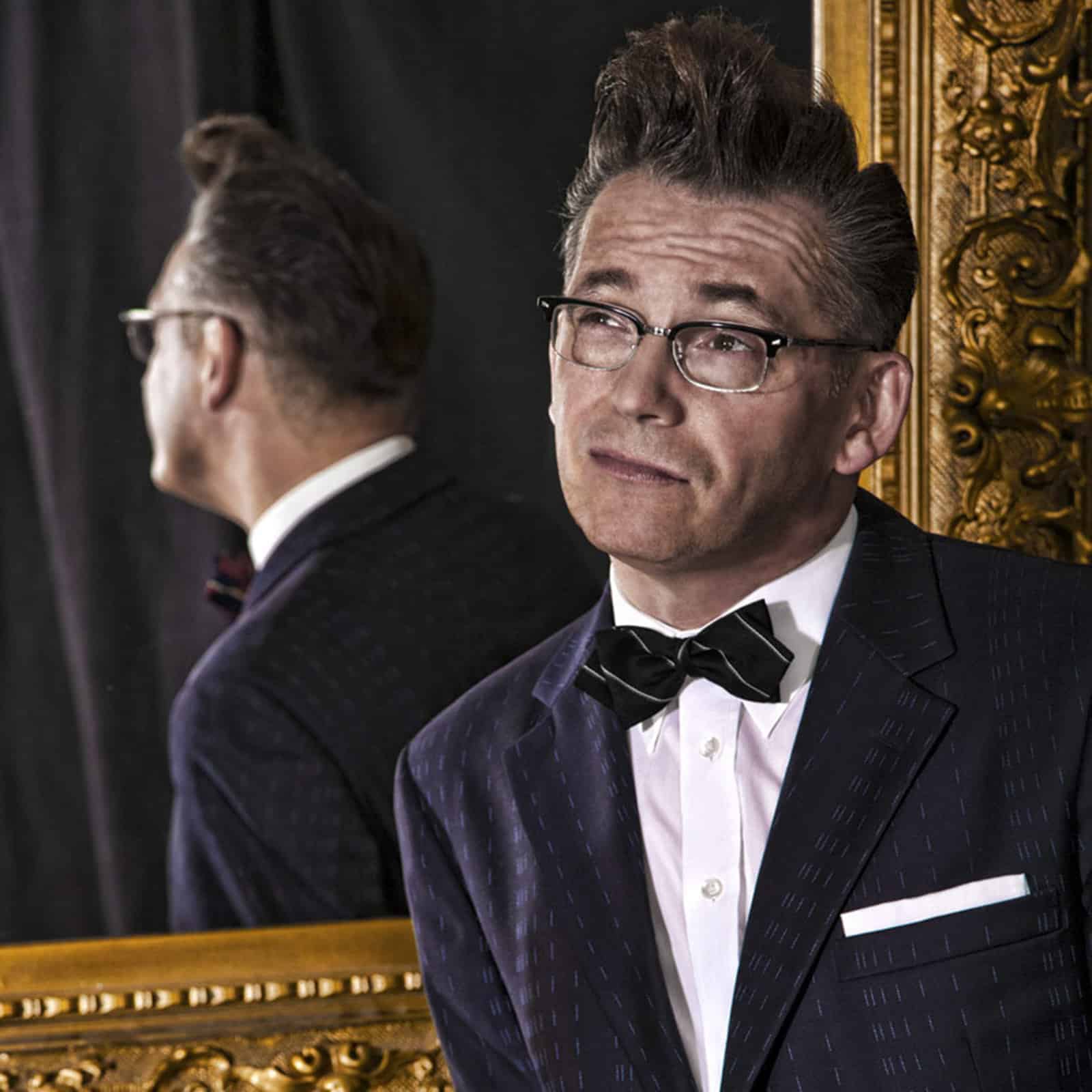 Ein Mann mit Brille, Fliege und dunklem Anzug mit weißem Einstecktuch steht vor einem verzierten, goldgerahmten Spiegel. Seine Haare sind hochgesteckt und nach hinten frisiert. Er blickt mit nachdenklichem Gesichtsausdruck leicht nach oben und sein Spiegelbild ist im Spiegel sichtbar. © Fotografie Tomas Rodriguez
