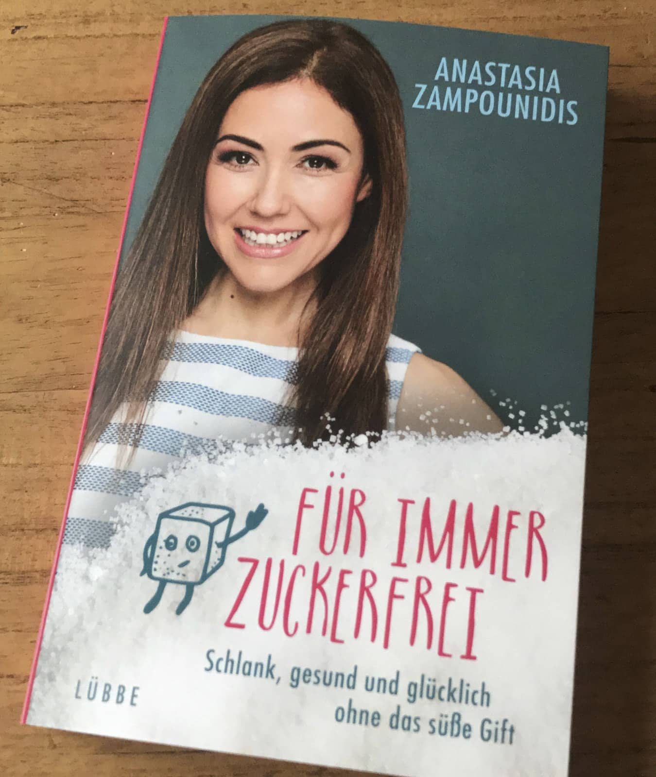 Auf einer Holzoberfläche liegt ein deutschsprachiges Buch mit dem Titel „Für immer zuckerfrei“ von Anastasia Zampounidis. Auf dem Cover ist eine lächelnde Frau mit langen dunklen Haaren und eine Comicfigur mit einem Zuckerwürfel abgebildet. Im Untertitel heißt es „Gesundheit und Glück ohne Zucker“. © Fotografie Tomas Rodriguez