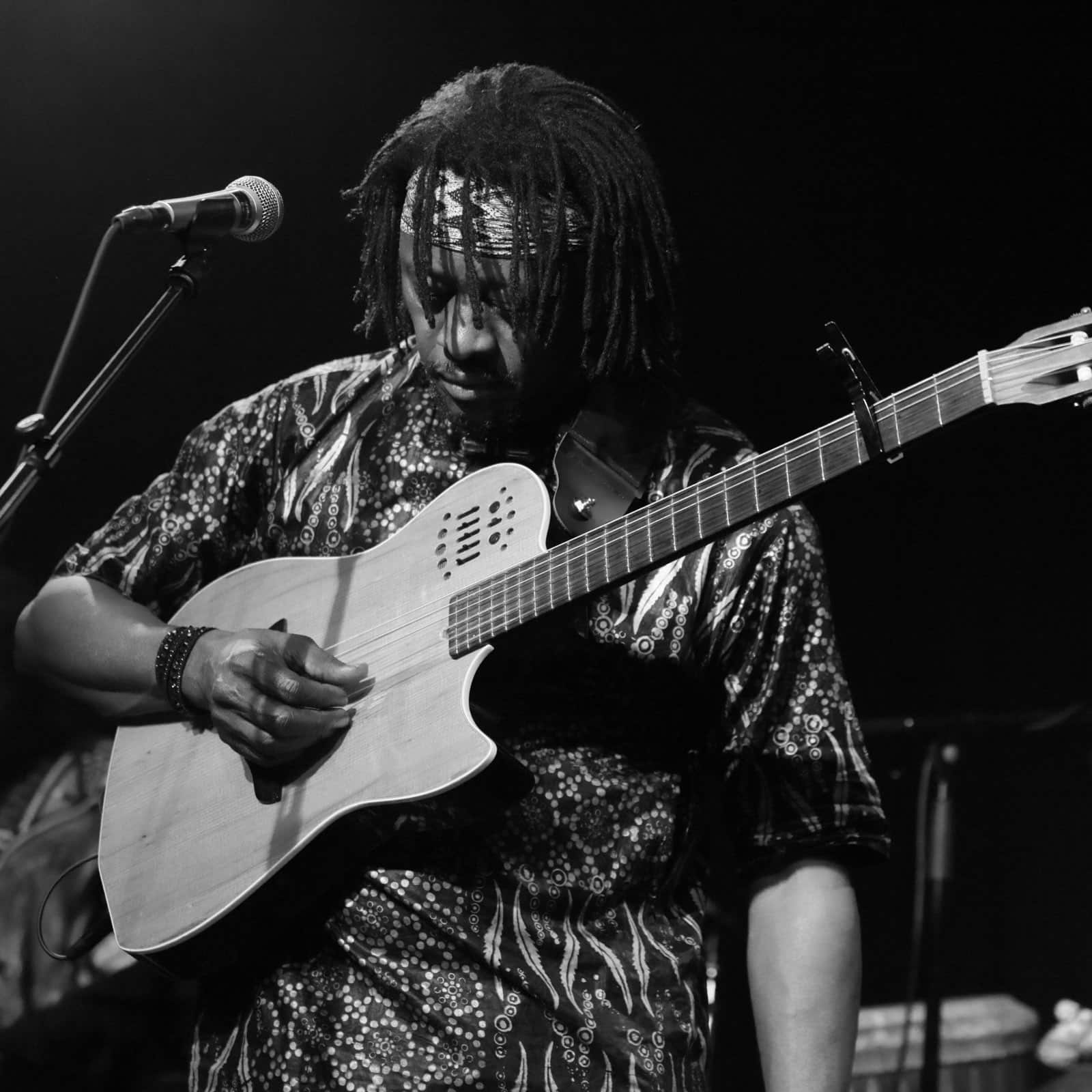 Ein Schwarzweißfoto eines Musikers, der auf der Bühne Akustikgitarre spielt. Der Musiker hat Dreadlocks und trägt ein gemustertes Hemd und ein Stirnband. In der Nähe steht ein Mikrofon auf einem Ständer, was auf einen Live-Auftritt schließen lässt. © Fotografie Tomas Rodriguez