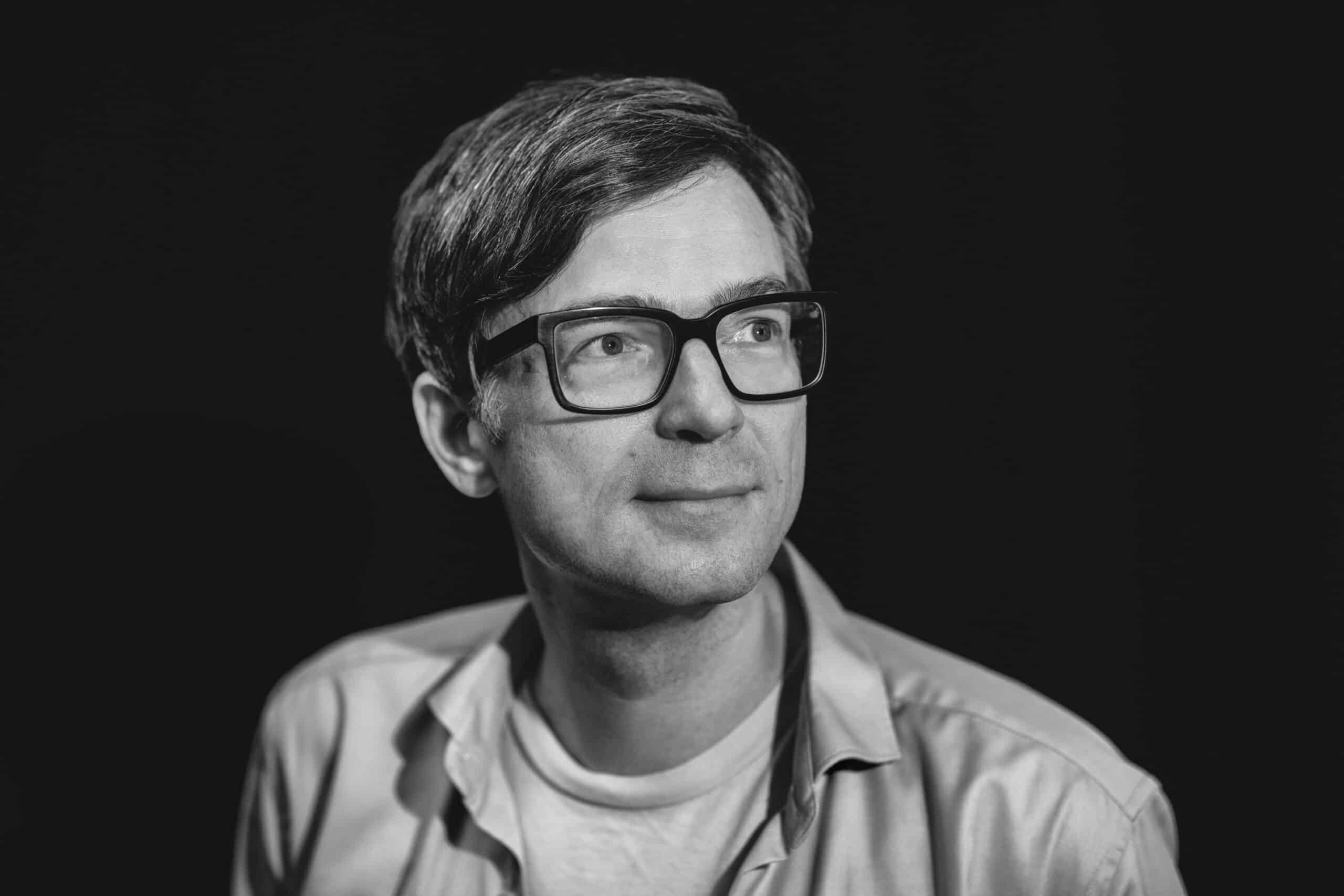 Ein Schwarzweißporträt einer Person mit kurzen, dunklen Haaren und Brille. Sie trägt ein helles Hemd und blickt mit nachdenklichem Gesichtsausdruck leicht nach links. Der Hintergrund ist dunkel, wodurch das Motiv hervorsticht. © Fotografie Tomas Rodriguez