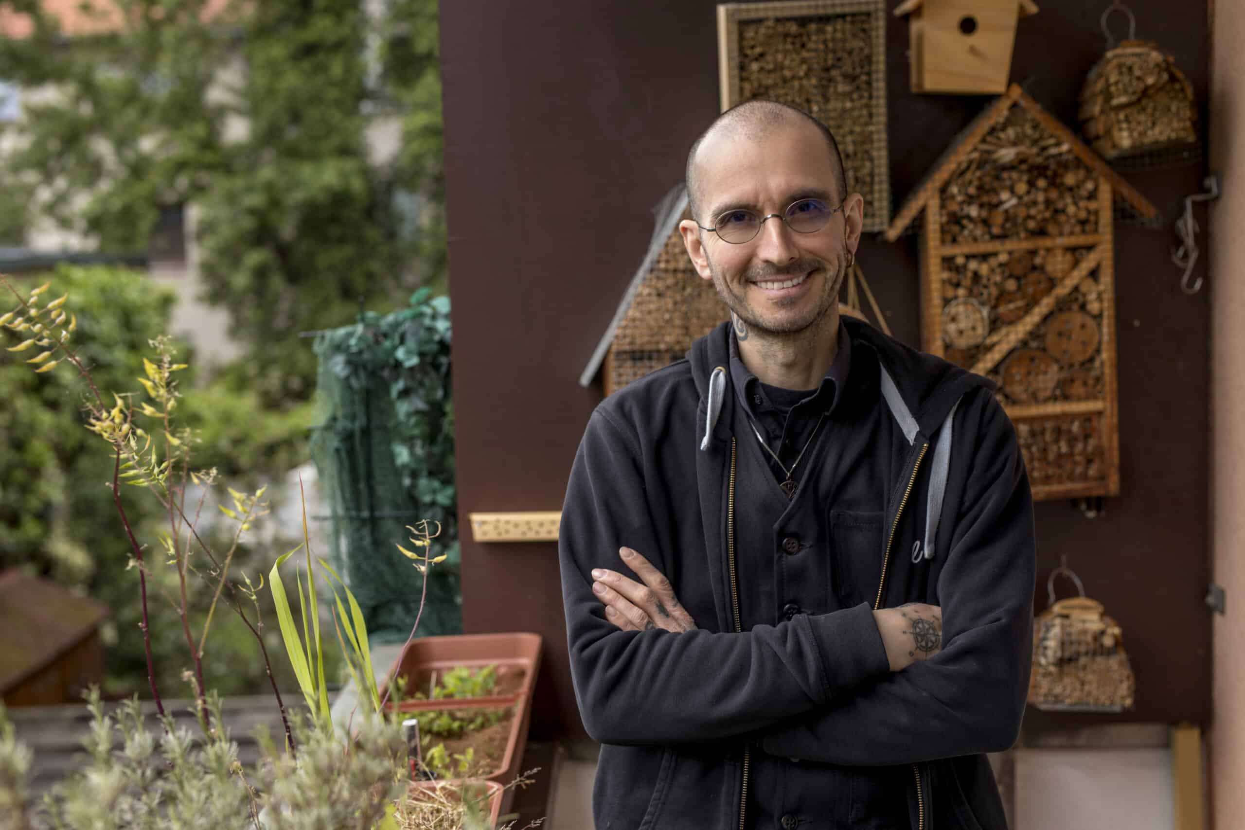Ein Mann mit Brille und schwarzer Jacke steht mit verschränkten Armen da und lächelt in die Kamera. Hinter ihm sind hölzerne Bienenhotels und Topfpflanzen zu sehen. Er steht auf einem Balkon, im Hintergrund sieht man Grün und Gebäude. © Fotografie Tomas Rodriguez
