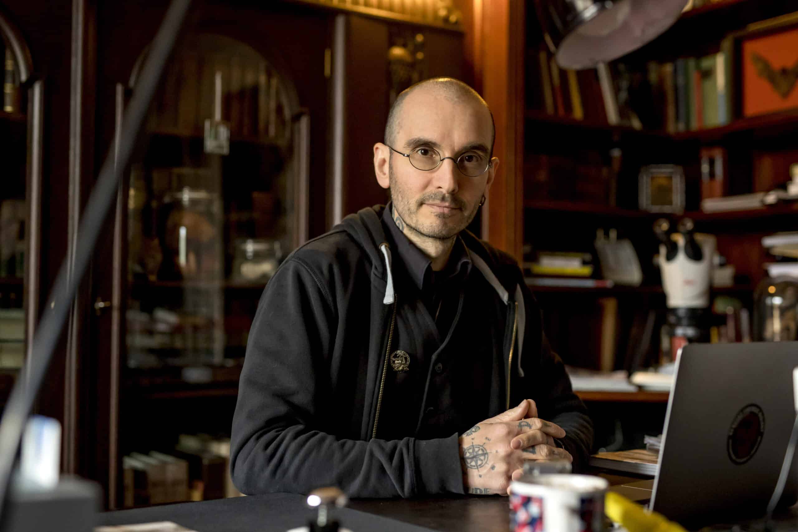 Eine Person mit Brille und schwarzem Kapuzenpullover sitzt an einem Schreibtisch in einem Raum mit Holzregalen voller Bücher und wissenschaftlicher Geräte. Sie hat ihre Hände auf dem Schreibtisch gefaltet und im Vordergrund sind ein Laptop, eine Tasse und verschiedene Gegenstände zu sehen. © Fotografie Tomas Rodriguez