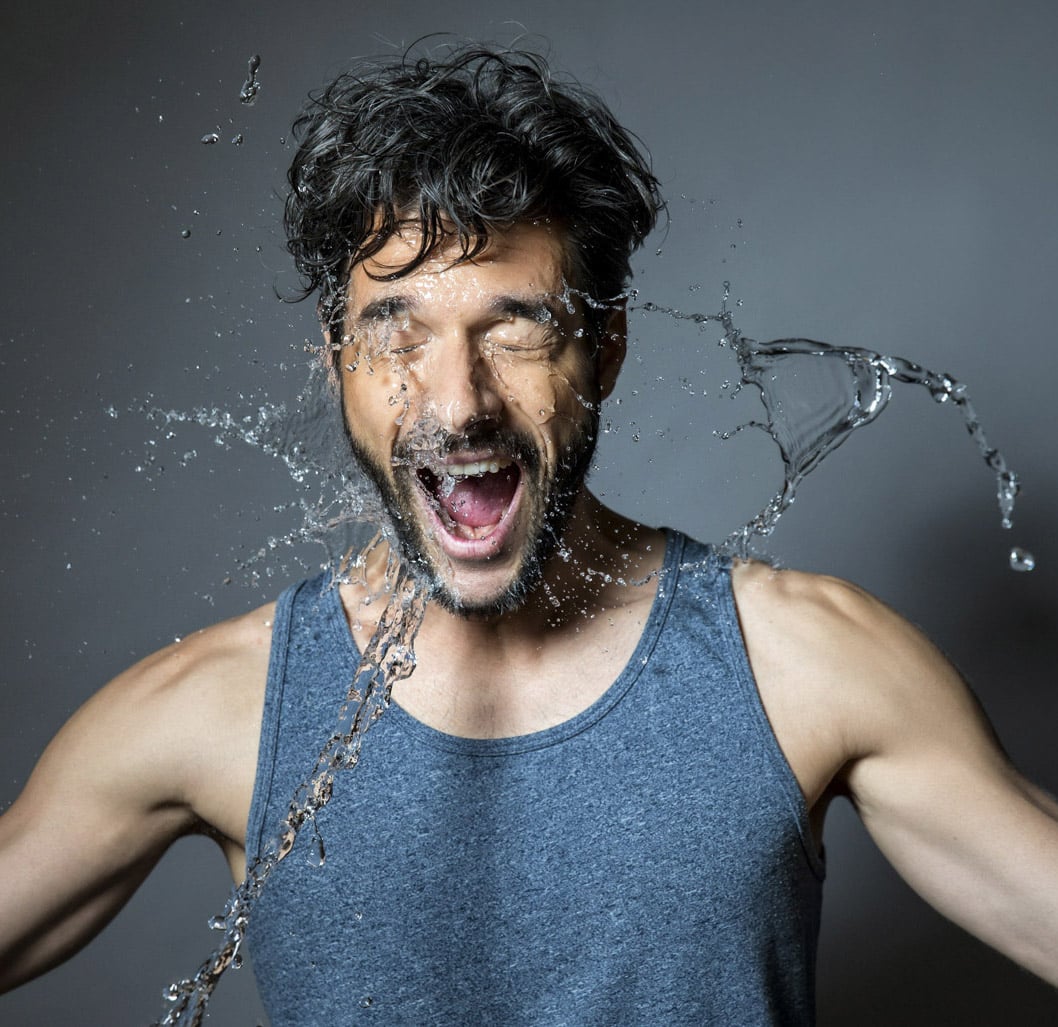 Ein Mann trägt ein graues Tanktop, hat kurzes, lockiges Haar und einen dichten Bart. Wasser spritzt ihm ins Gesicht, während er die Augen geschlossen hat und sein Mund offen ist, als würde er schreien oder auf das Spritzwasser reagieren. Der Hintergrund ist schlicht grau. © Fotografie Tomas Rodriguez