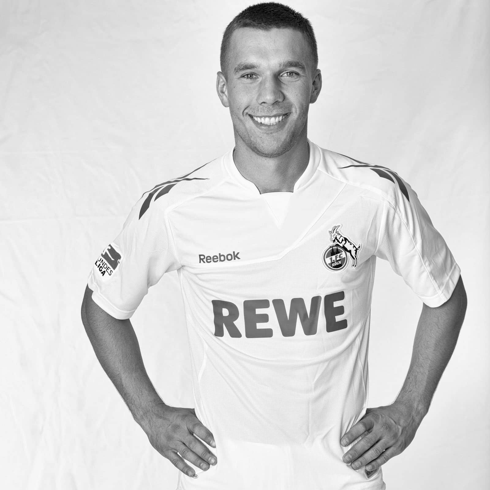 Schwarzweißbild eines lächelnden Fußballspielers, der mit den Händen in den Hüften steht. Er trägt ein Reebok-Trikot mit dem Wort „REWE“ auf der Vorderseite und einem Wappen auf der linken Brust. Der Hintergrund ist schlicht und weiß. © Fotografie Tomas Rodriguez