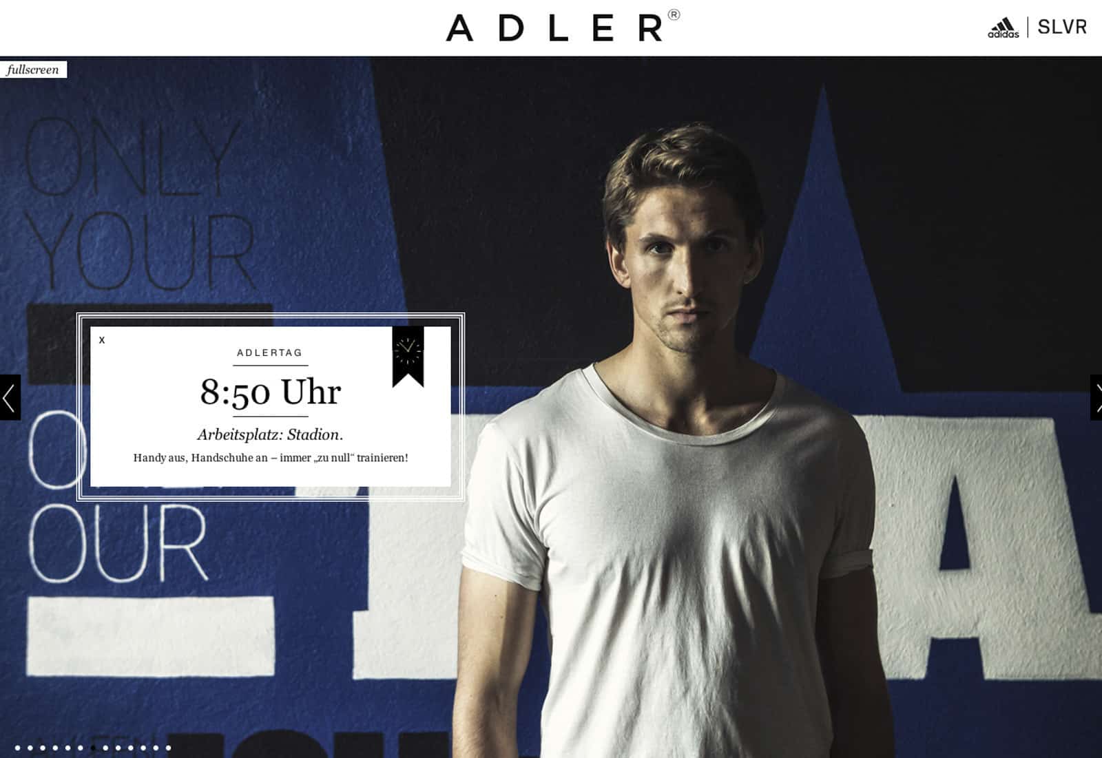 Ein Mann in einem weißen T-Shirt steht vor einer blau-schwarzen Wand mit Text. Der darüberliegende Text lautet auf Deutsch: „ADLERTAG 8:50 Uhr Arbeitsplatz: Stadion“. In den oberen Ecken des Bildes sind Adidas- und SLVR-Logos zu sehen. © Fotografie Tomas Rodriguez