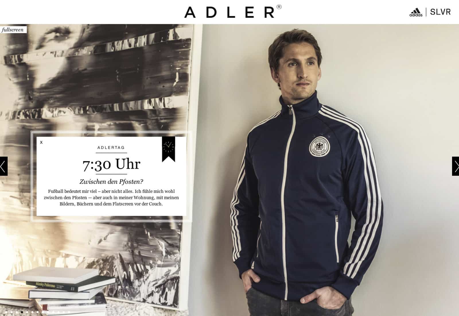 Ein junger Mann steht in einem modernen Wohnzimmer. Er trägt eine marineblaue Adidas-Trainingsjacke mit weißen Streifen und einem deutschen Emblem. Im Hintergrund sind ein Schwarzweißfoto, Bücher und Dekorationsgegenstände auf Regalen zu sehen. Der Text lautet „7:30 Uhr Zwischen den Pfosten?“. © Fotografie Tomas Rodriguez