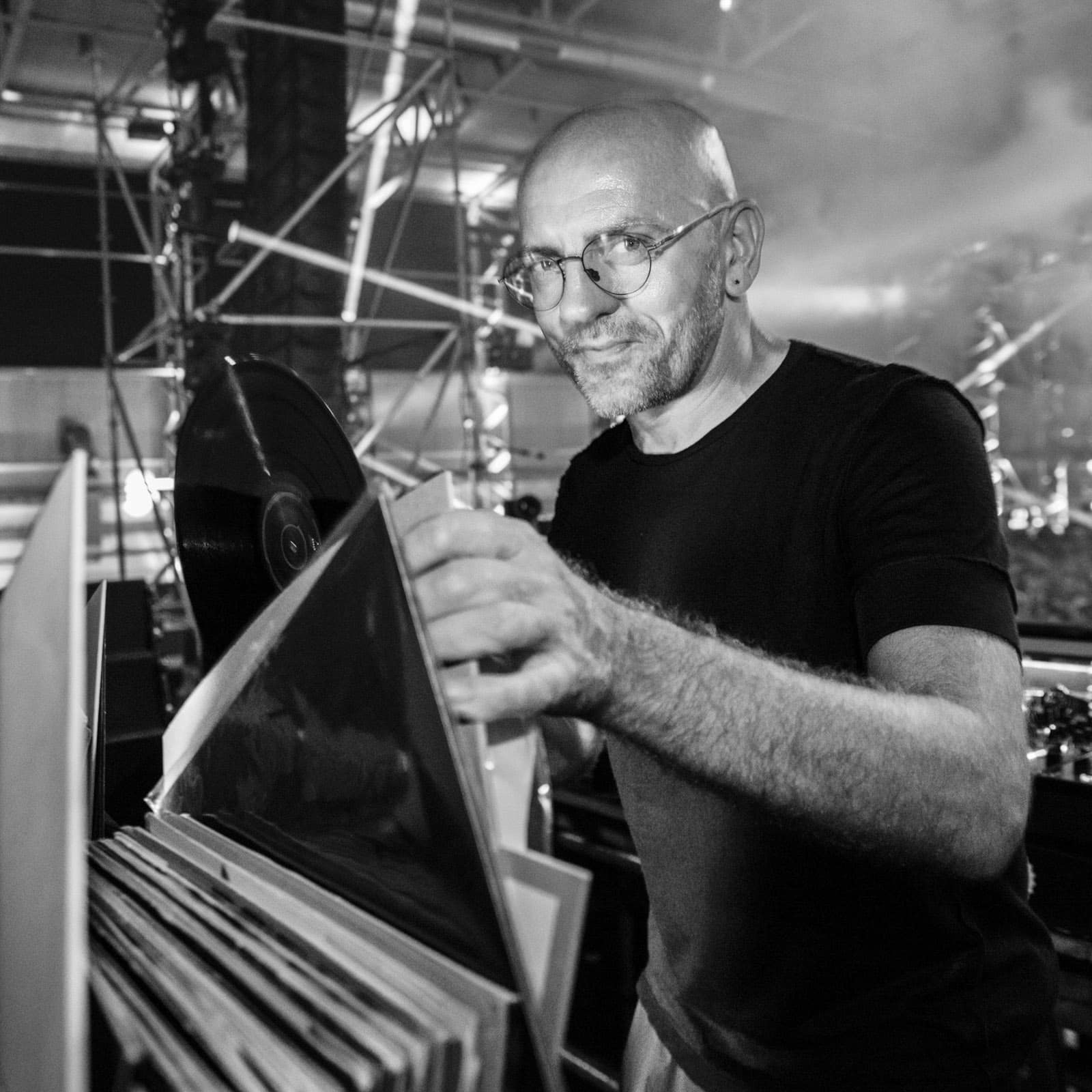 Ein Mann mit Brille und Bart sortiert bei einer Open-Air-Veranstaltung oder einem Konzert Schallplatten. Er trägt ein schwarzes T-Shirt und scheint DJ zu sein. Im Hintergrund sind Bühnentechnik und Beleuchtung zu sehen, was auf eine laufende Aufführung schließen lässt. Schwarzweißfoto. © Fotografie Tomas Rodriguez