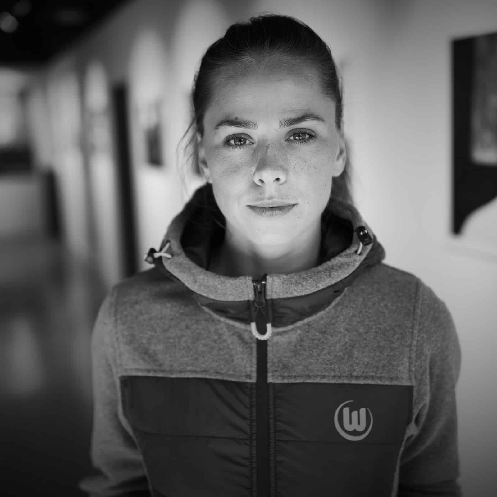 Eine Frau mit ernster Miene steht in einem Flur und trägt eine Jacke mit einem Logo darauf. Die Umgebung ist mit sanftem, fokussiertem Licht beleuchtet und im Hintergrund sind gerahmte Bilder oder Kunstwerke an den Wänden zu sehen. Das Bild ist schwarzweiß. © Fotografie Tomas Rodriguez
