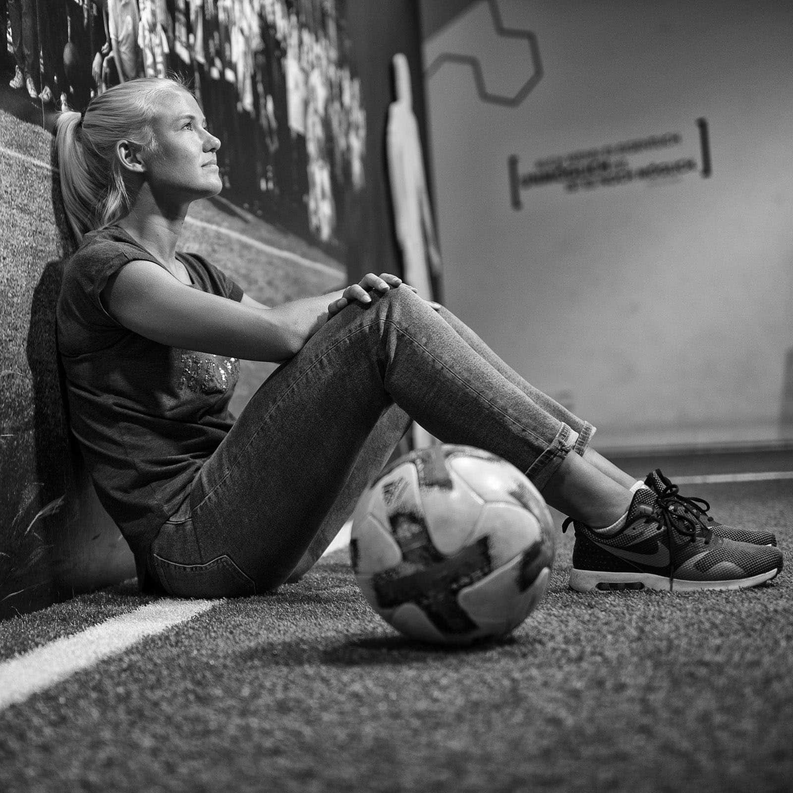 Eine Frau mit zurückgebundenen langen blonden Haaren sitzt mit dem Rücken an einer Wand auf dem Boden, die Arme auf den Knien ruhend, und blickt nachdenklich nach oben. Vor ihr liegt ein Fußball auf dem Boden. Die Szenerie scheint eine Fußballhalle zu sein. Schwarzweißfoto. © Fotografie Tomas Rodriguez