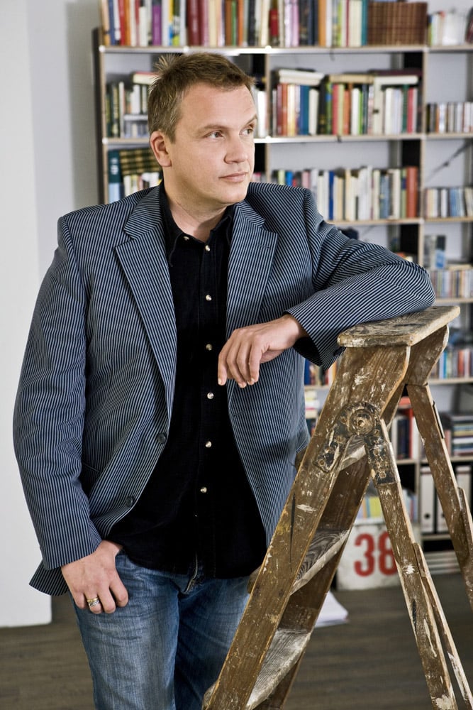 Ein Mann lehnt in einer Bibliothek auf einer Holzleiter. Er trägt einen Blazer mit feinen blau-weißen Streifen, ein schwarzes Hemd und Jeans. Im Hintergrund sind Bücherregale voller Bücher zu sehen. © Fotografie Tomas Rodriguez