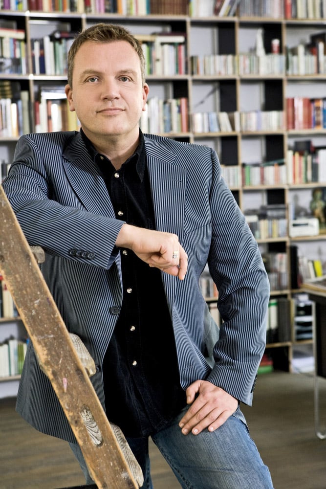 Ein Mann in Nadelstreifenblazer und Jeans lehnt in einer Bibliothek an einer Holzleiter. Er hat kurzes, helles Haar und blickt mit neutralem Gesichtsausdruck in die Kamera. Im Hintergrund sind Regale voller Bücher zu sehen. © Fotografie Tomas Rodriguez