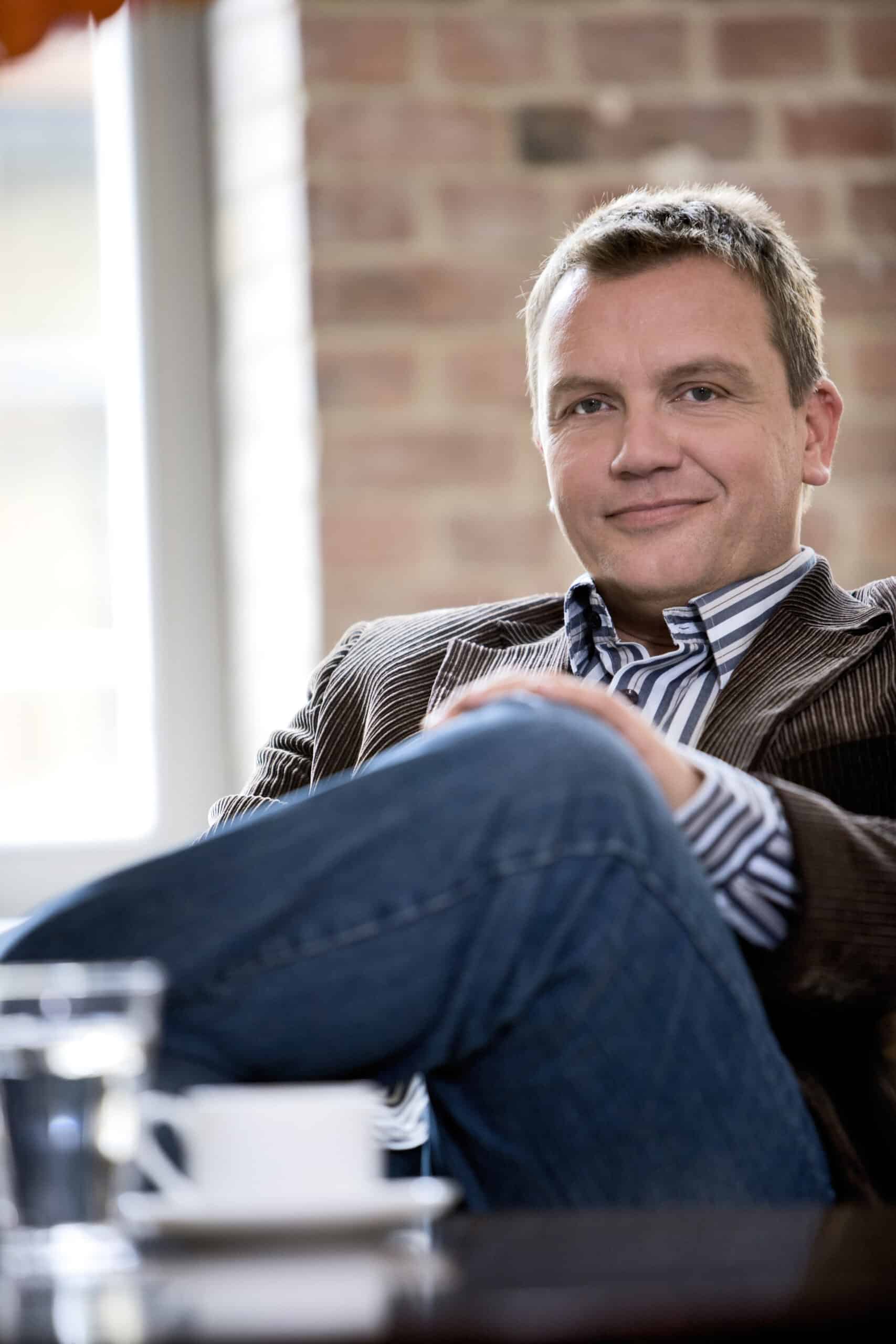 Ein Mann mit kurzen Haaren sitzt bequem mit gekreuzten Beinen da und trägt einen dunkel gestreiften Blazer über einem gestreiften Hemd und Jeans. Er befindet sich in einer entspannten Umgebung mit einer Backsteinmauer und einem Fenster im Hintergrund und einem Tisch mit einer Tasse und einem Glas Wasser im Vordergrund. © Fotografie Tomas Rodriguez