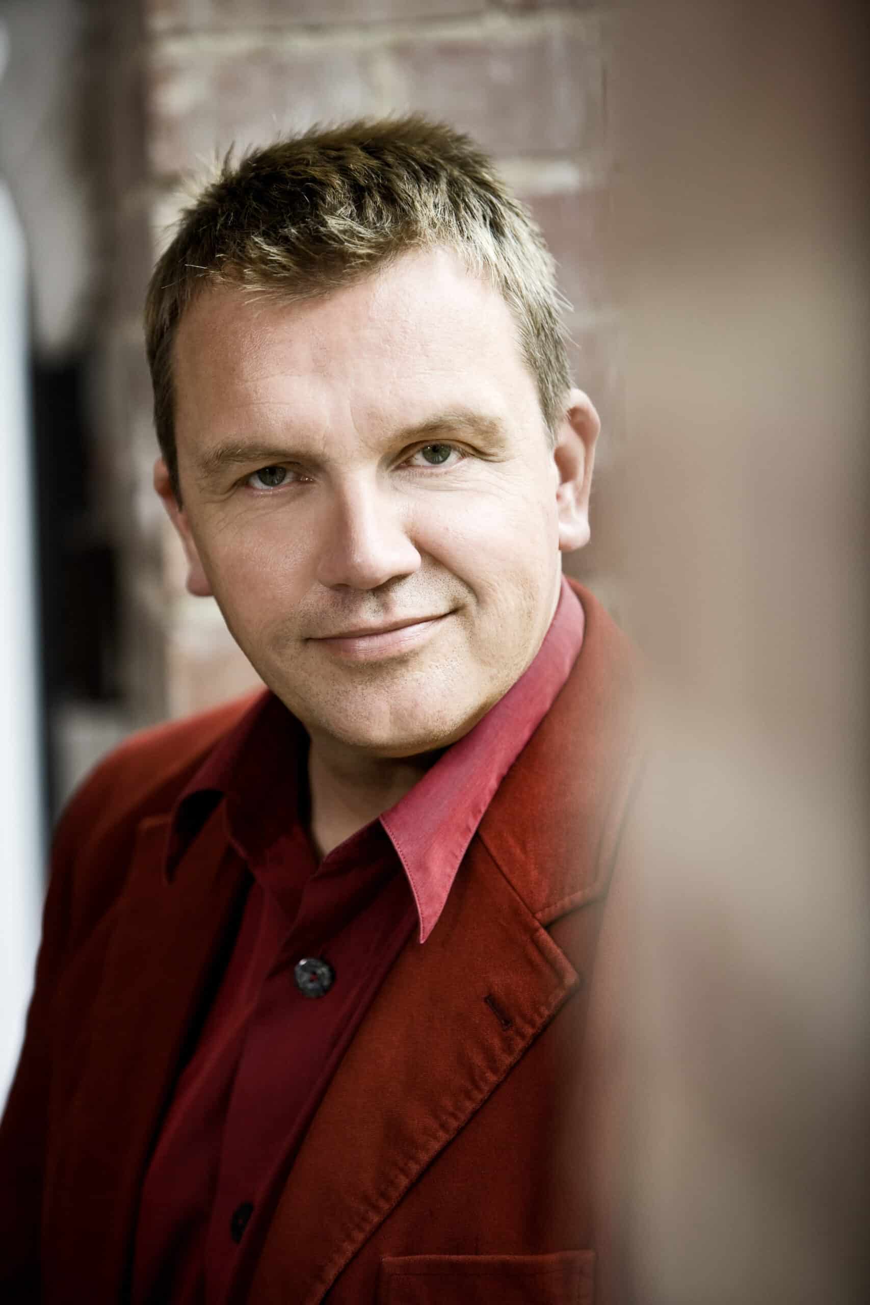 Ein Mann mit kurzen blonden Haaren lächelt leicht. Er trägt ein rotes Oberhemd und einen roten Blazer. Der Hintergrund ist verschwommen, aber es scheint sich um eine Außenkulisse mit einer Backsteinmauer zu handeln. Er lehnt sich leicht nach links. © Fotografie Tomas Rodriguez