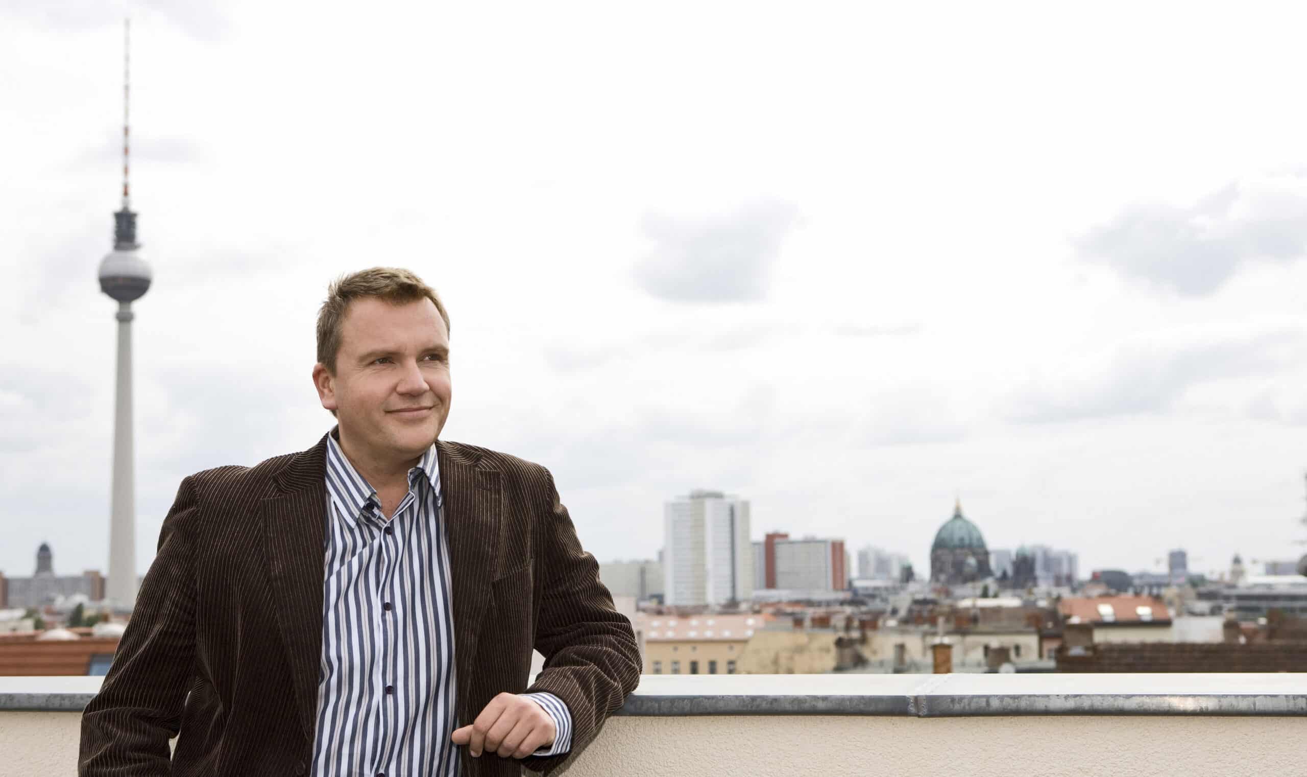 Eine Person in brauner Jacke und gestreiftem Hemd steht auf einem Dach mit einer Stadtlandschaft im Hintergrund. Die Skyline umfasst einen markanten Fernsehturm und mehrere Gebäude mit Kuppeln. Die Person blickt mit einem leichten Lächeln in die Ferne. © Fotografie Tomas Rodriguez