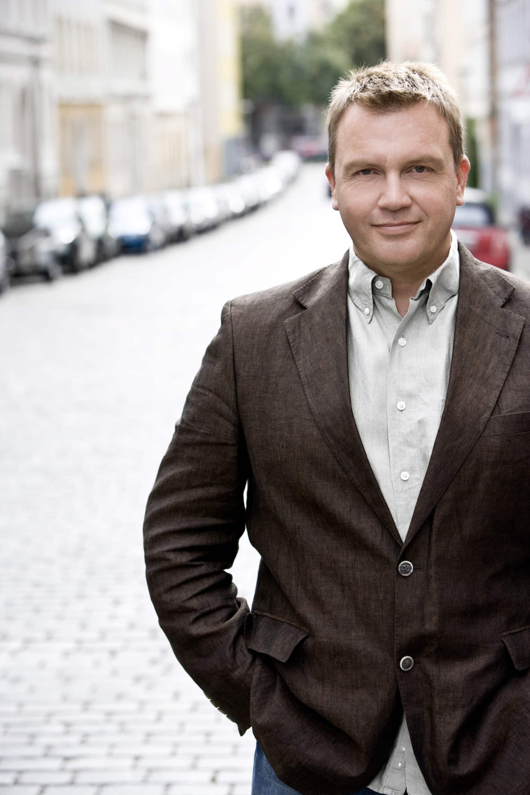 Ein Mann mit kurzen blonden Haaren steht auf einer Kopfsteinpflasterstraße, die von geparkten Autos gesäumt ist. Er trägt einen braunen Blazer über einem hellgrauen Hemd und hat die Hände lässig in den Taschen. Der Hintergrund ist eine städtische Umgebung mit Gebäuden auf beiden Seiten. © Fotografie Tomas Rodriguez