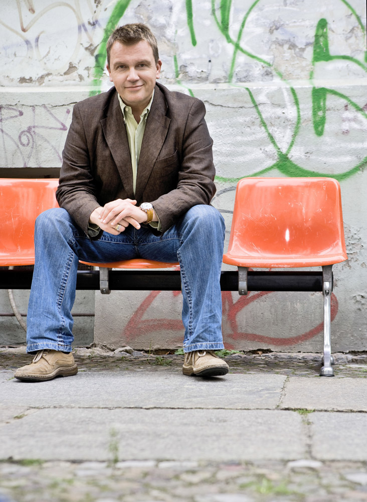 Eine Person sitzt auf einem orangefarbenen Stuhl im Stadionstil im Freien vor einer mit Graffiti bedeckten Wand. Sie ist leger gekleidet in einem braunen Blazer, einem grünen Hemd, blauen Jeans und hellbraunen Schuhen und faltet die Hände, während sie mit einem leichten Lächeln in die Kamera blickt. © Fotografie Tomas Rodriguez