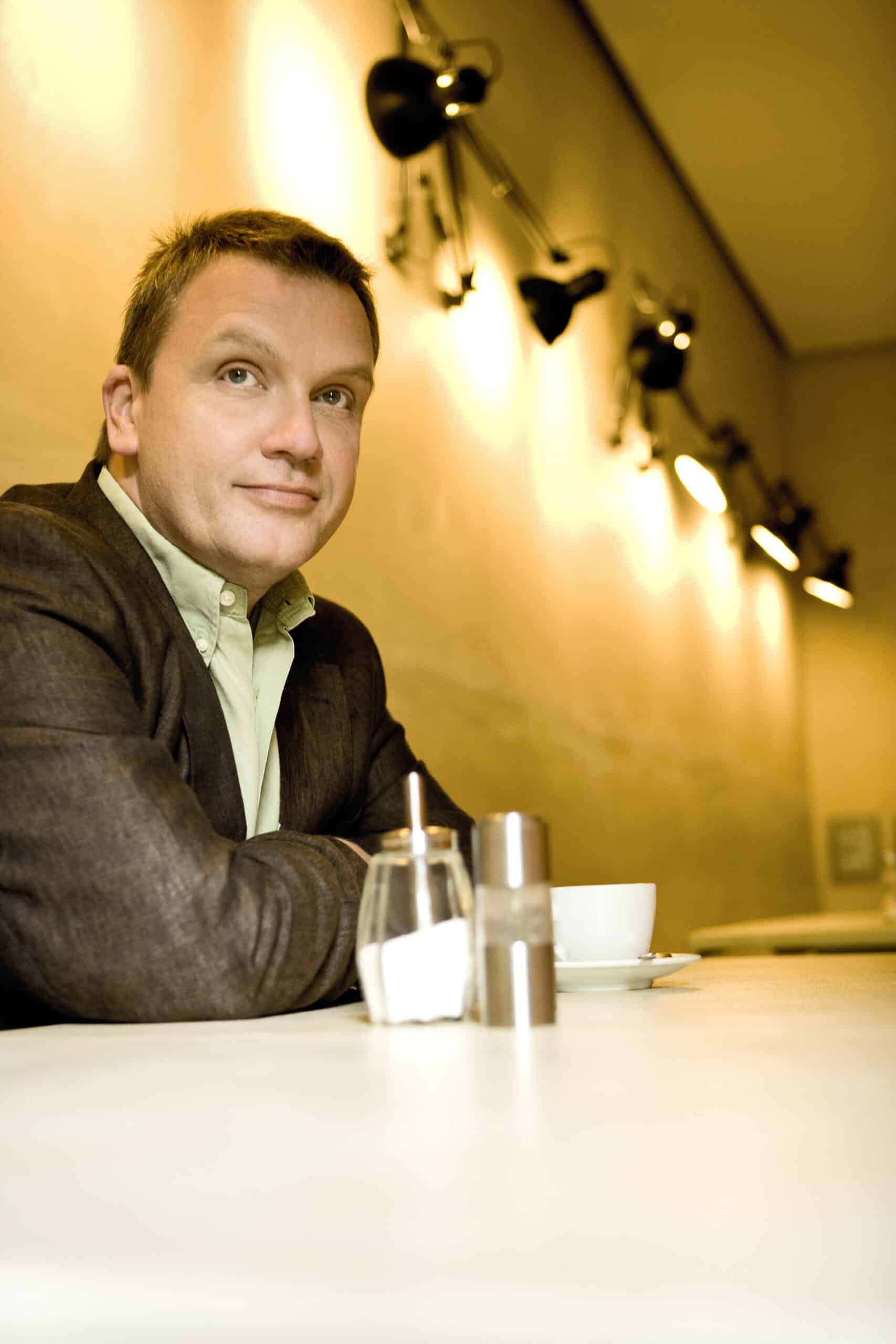 Ein Mann sitzt an einem Tisch in einem gut beleuchteten Café. Er trägt einen braunen Blazer über einem hellen Hemd und blickt mit einem leichten Lächeln zur Seite. Auf dem Tisch vor ihm stehen ein Zuckerspender, ein Salzstreuer und eine weiße Kaffeetasse. © Fotografie Tomas Rodriguez