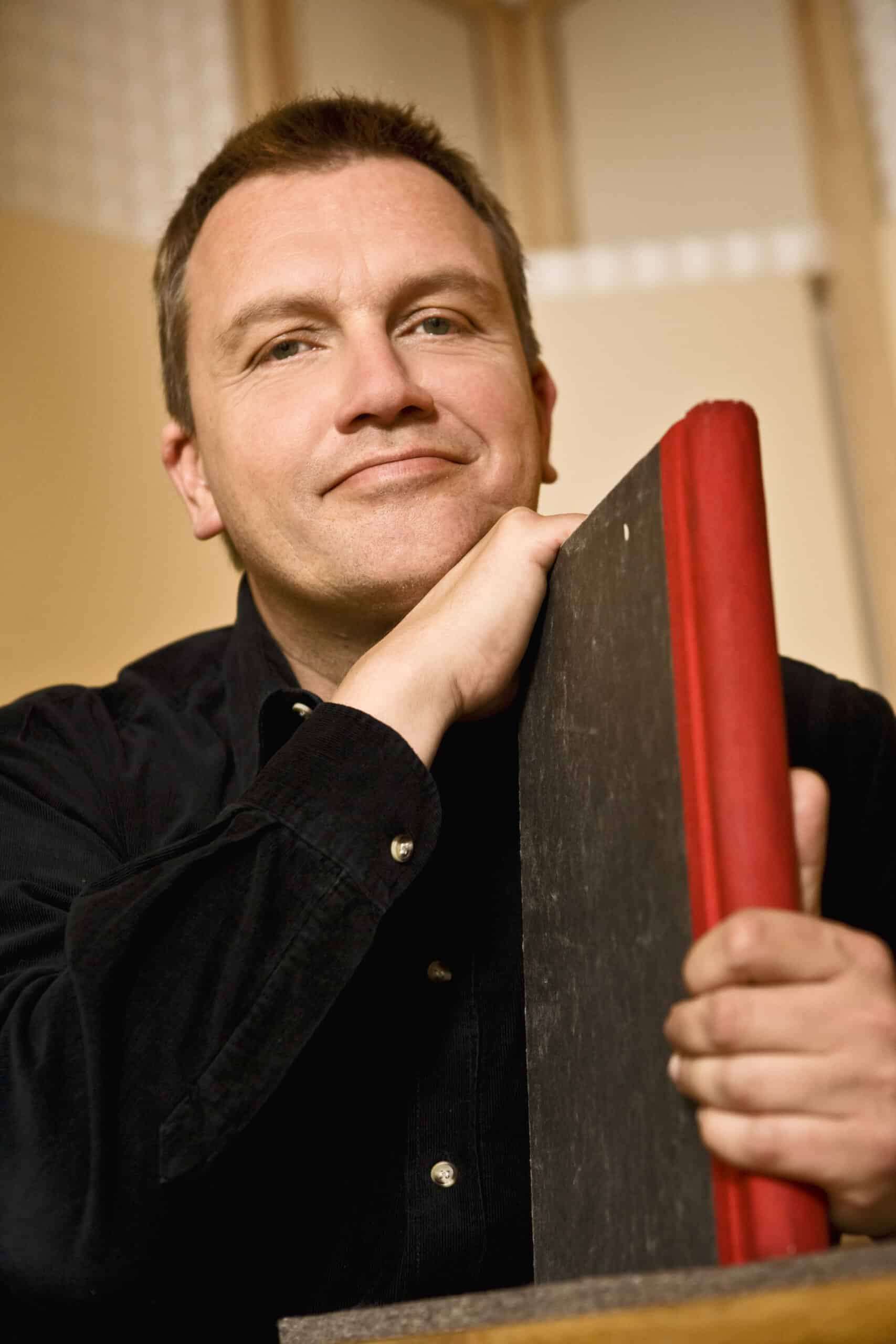 Eine Person mit kurzen Haaren stützt ihr Kinn auf ihre Hand, die auf einem großen, dunklen Buch mit rotem Rücken ruht. Sie trägt ein schwarzes Hemd mit Knöpfen und lächelt leicht, während sie in die Kamera blickt. Der Hintergrund ist leicht unscharf. © Fotografie Tomas Rodriguez