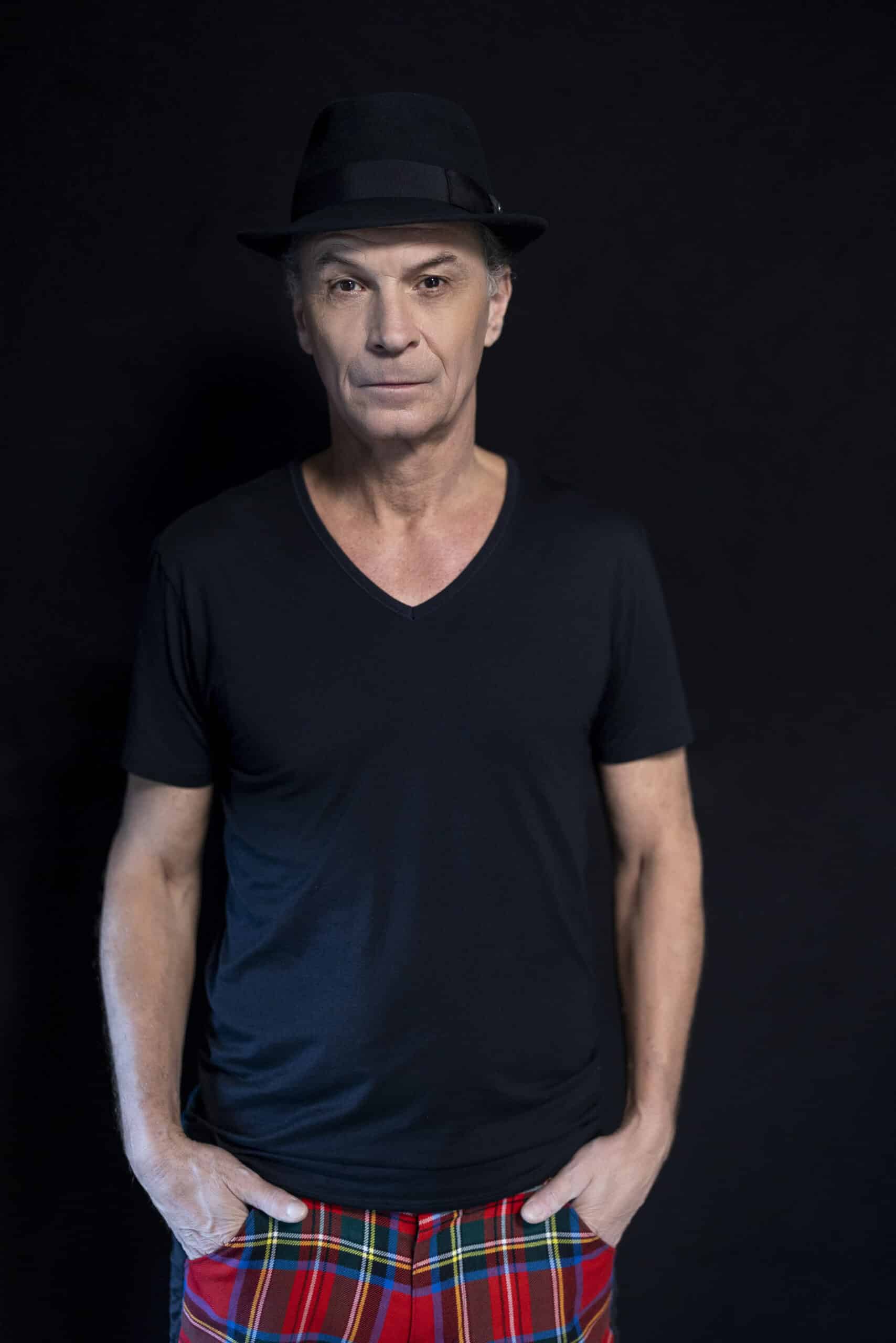 Ein Mann mittleren Alters trägt ein schwarzes T-Shirt mit V-Ausschnitt, einen schwarzen Fedora-Hut und rot karierte Hosen. Er steht mit den Händen in den Taschen vor einem dunklen Hintergrund und blickt direkt in die Kamera. © Fotografie Tomas Rodriguez