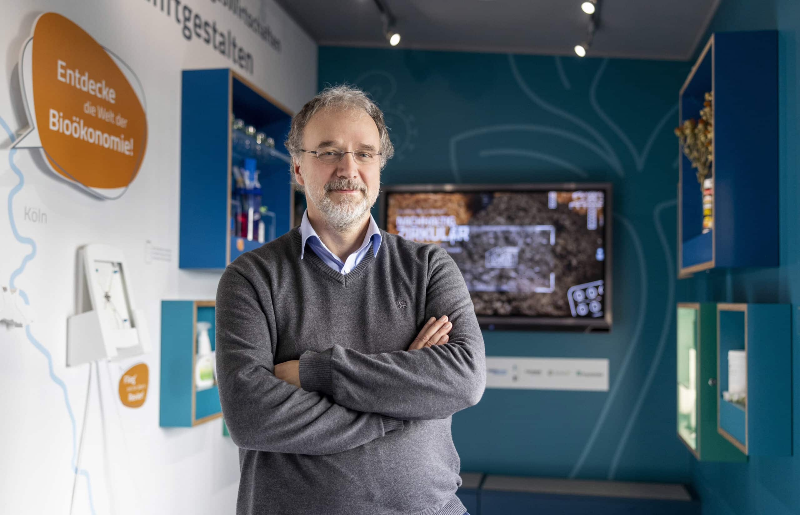 Ein Mann mit grauen Haaren und Brille steht mit verschränkten Armen in einem modernen Ausstellungsraum. Er trägt einen grauen Pullover über einem Hemd. Hinter ihm befindet sich eine Wand mit Informationstafeln, einem Videobildschirm und verschiedenen Exponaten. © Fotografie Tomas Rodriguez