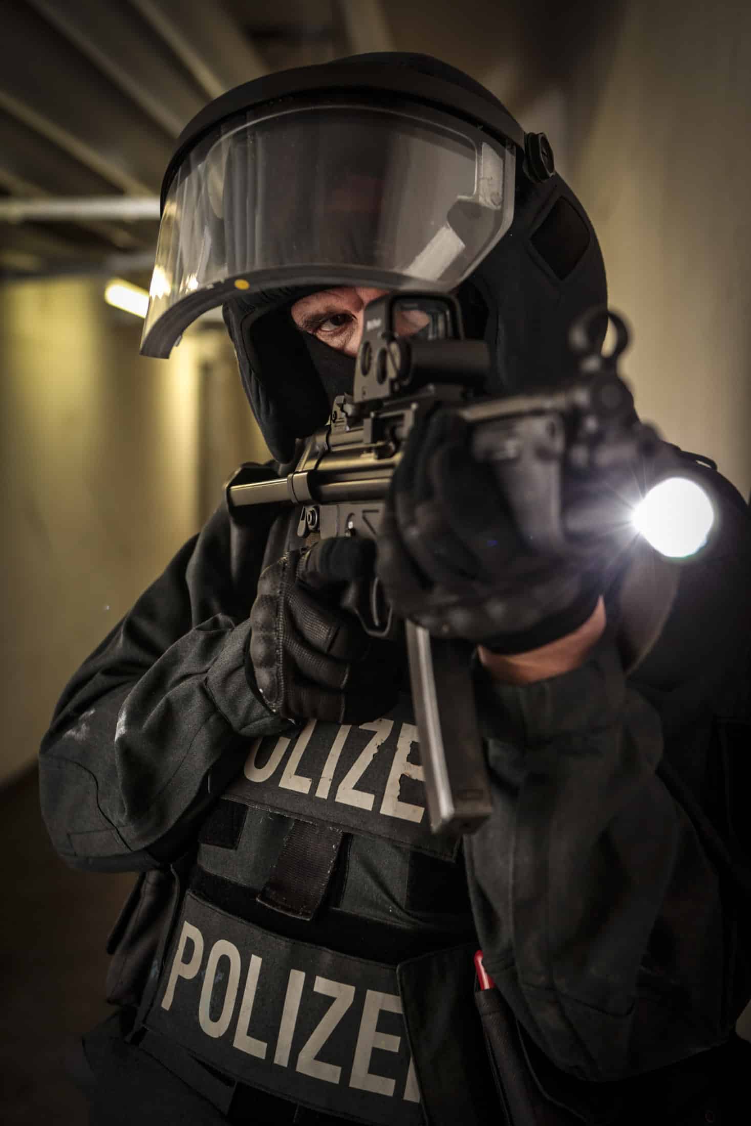 Ein Polizist in taktischer Ausrüstung mit der Aufschrift „Polizei“ zielt mit einem Gewehr, an dem eine Taschenlampe befestigt ist. Der Beamte trägt einen Helm mit Visier und Handschuhe und scheint sich in einem schwach beleuchteten Korridor oder Flur zu befinden. © Fotografie Tomas Rodriguez