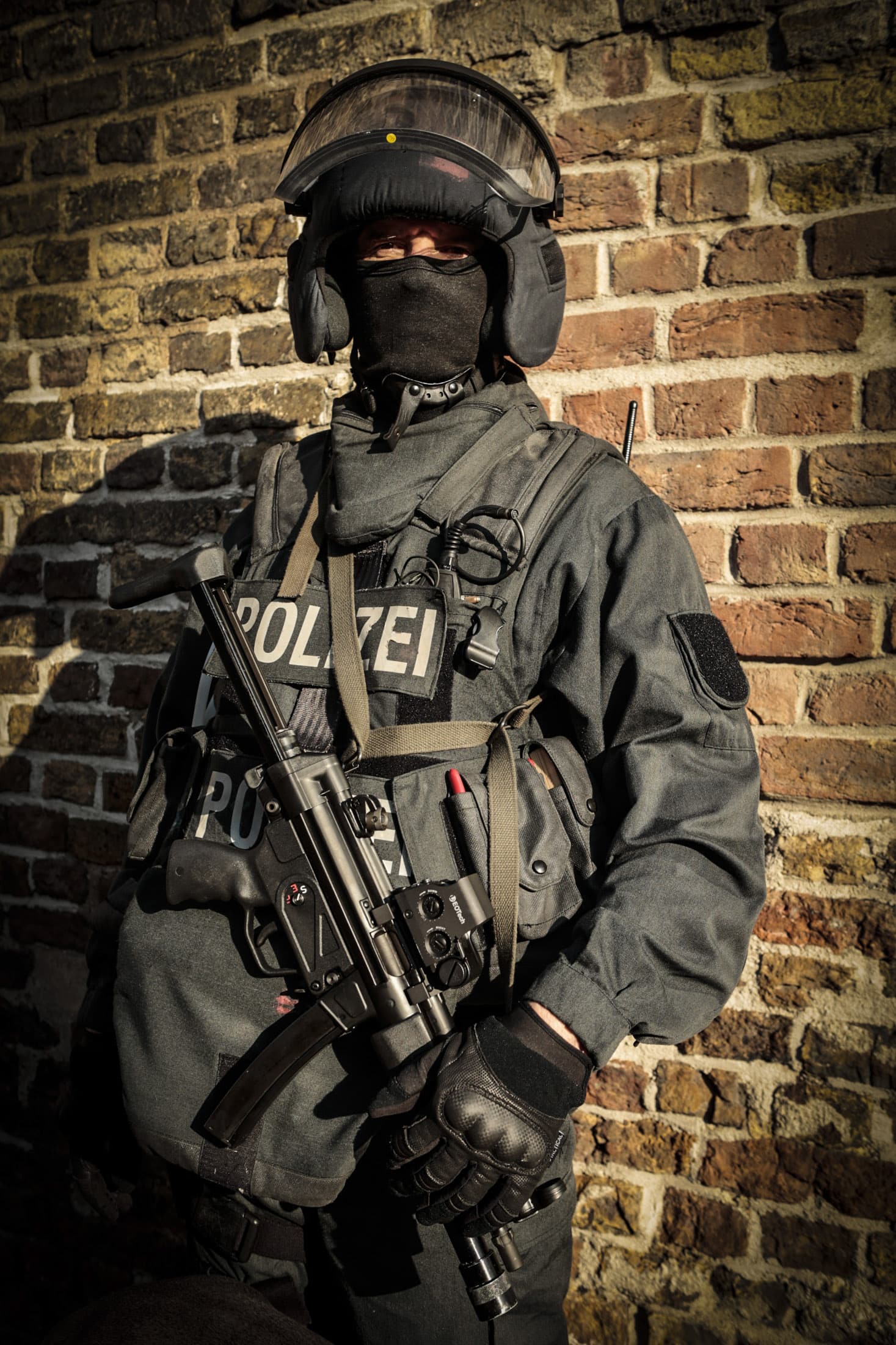 Ein taktischer Polizist in voller Montur steht vor einer Backsteinmauer. Er trägt einen Helm, eine Gesichtsmaske und Handschuhe und ist mit einer Schusswaffe, einem Funkgerät und verschiedener taktischer Ausrüstung ausgestattet. Auf seiner Weste ist das Wort „Polizei“ zu sehen. Sonnenlicht wirft Schatten auf die Mauer. © Fotografie Tomas Rodriguez