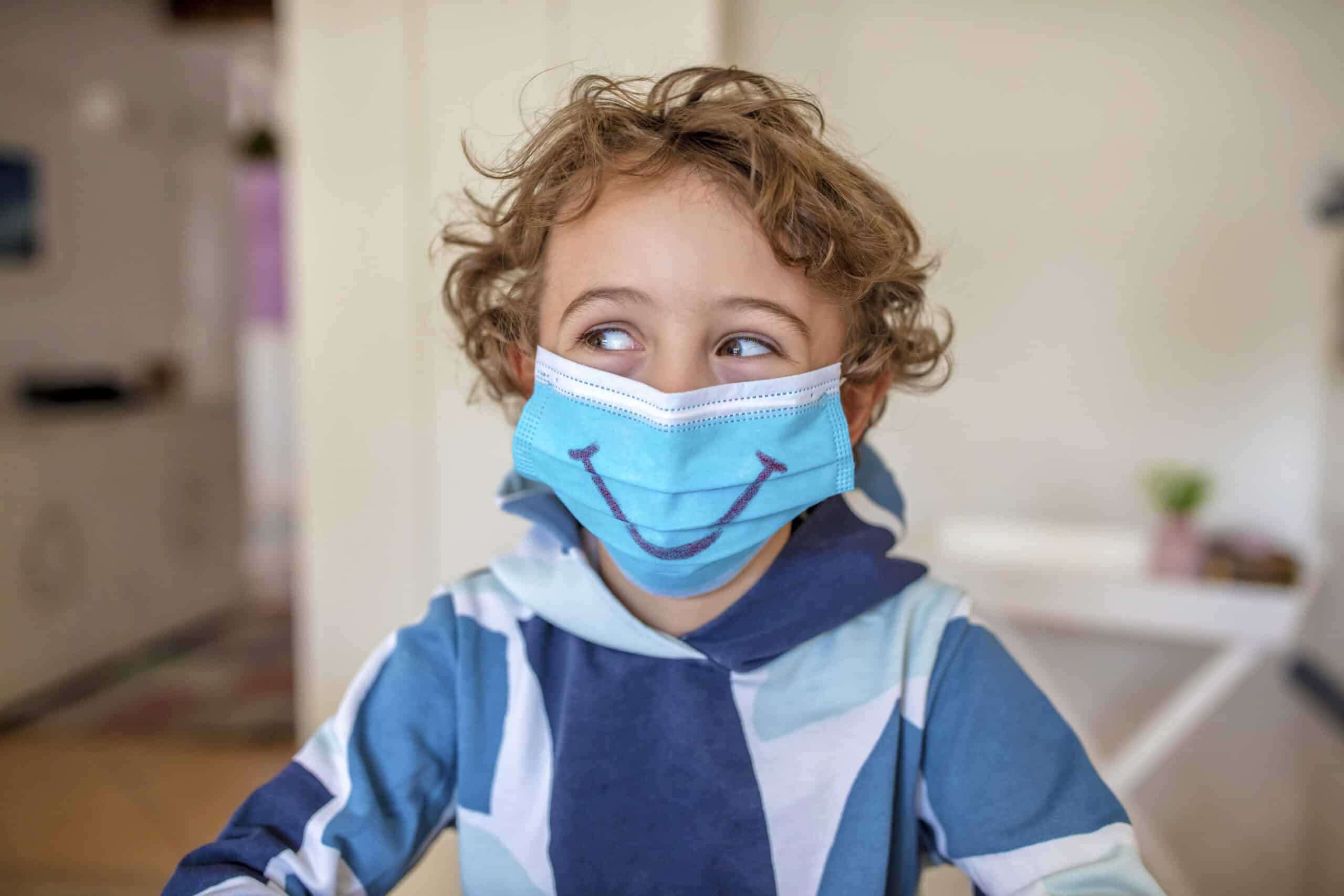Ein kleines Kind mit lockigem Haar trägt eine blaue Gesichtsmaske mit einem darauf gemalten Smiley. Das Kind blickt zur Seite und trägt einen blau-weiß gemusterten Kapuzenpullover. Der Hintergrund ist eine sanft fokussierte Innenkulisse. © Fotografie Tomas Rodriguez