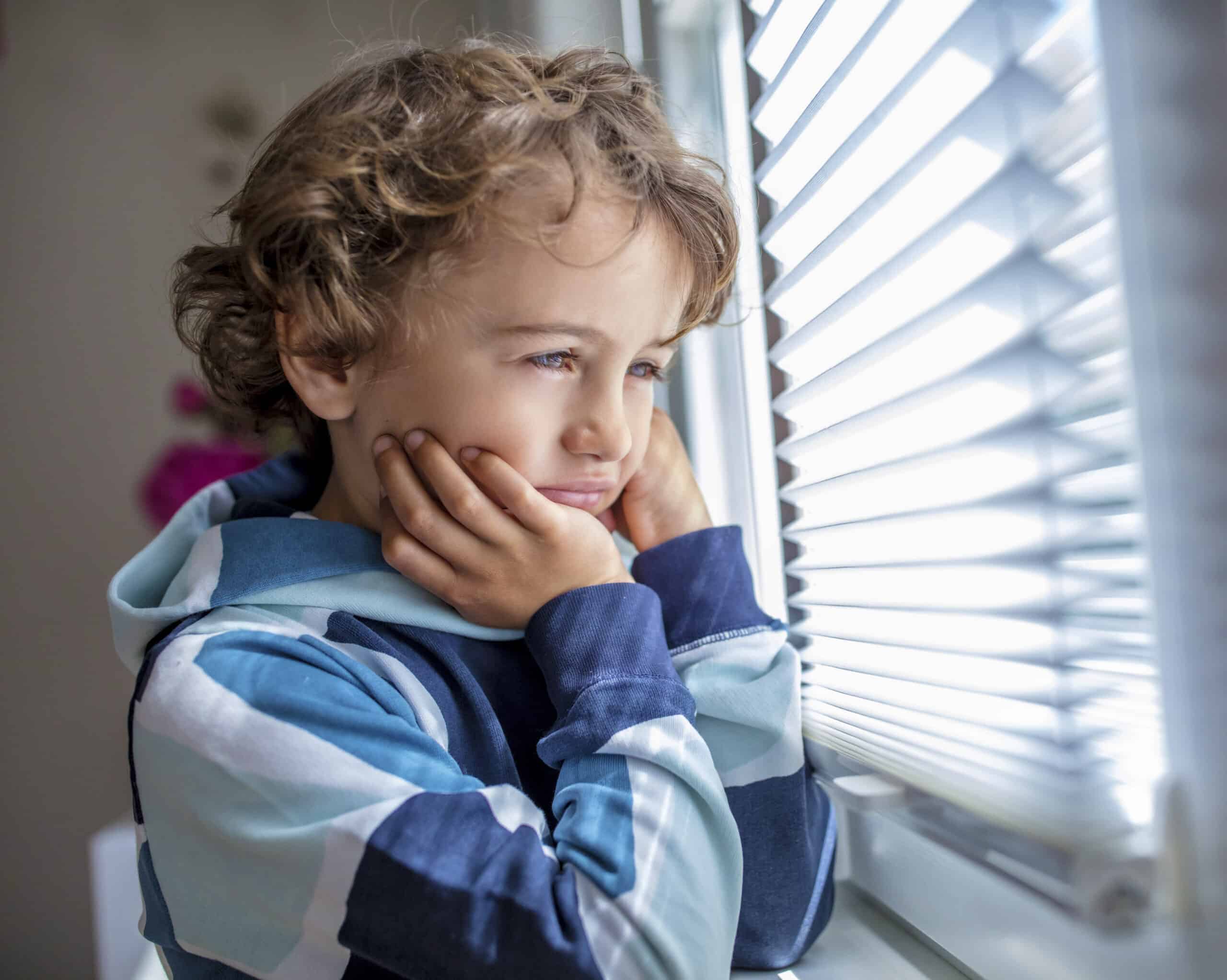 Ein kleines Kind mit lockigem Haar schaut nachdenklich aus einem Fenster mit geschlossenen Jalousien und stützt sein Kinn auf die Hände. Das Kind trägt einen gestreiften Kapuzenpullover und scheint in Gedanken versunken oder tagträumend zu sein. Licht fällt durch die Jalousien. © Fotografie Tomas Rodriguez