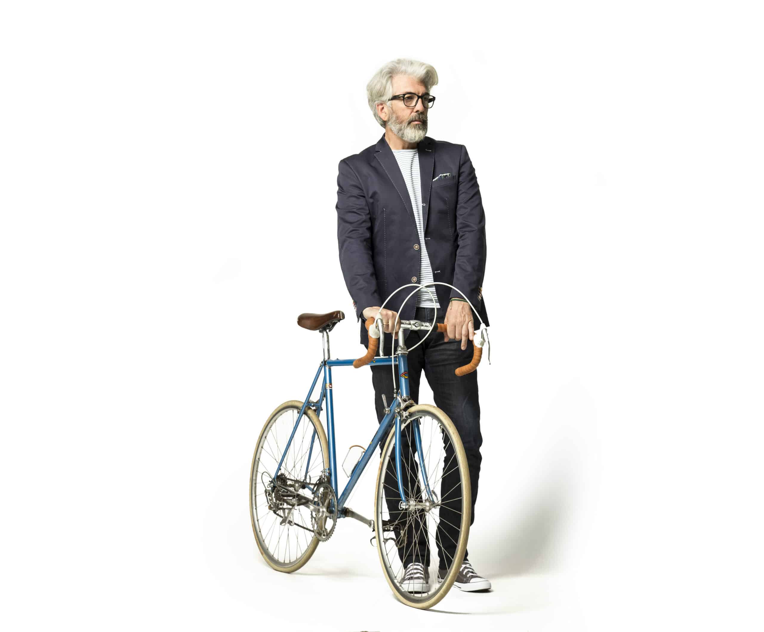 Ein vornehmer älterer Mann mit grauem Haar, Brille und Bart steht da und hält ein blaues Vintage-Fahrrad mit hellbraunem Lenker. Er trägt einen marineblauen Blazer, ein gestreiftes Hemd, Jeans und Turnschuhe. Der Hintergrund ist weiß. © Fotografie Tomas Rodriguez