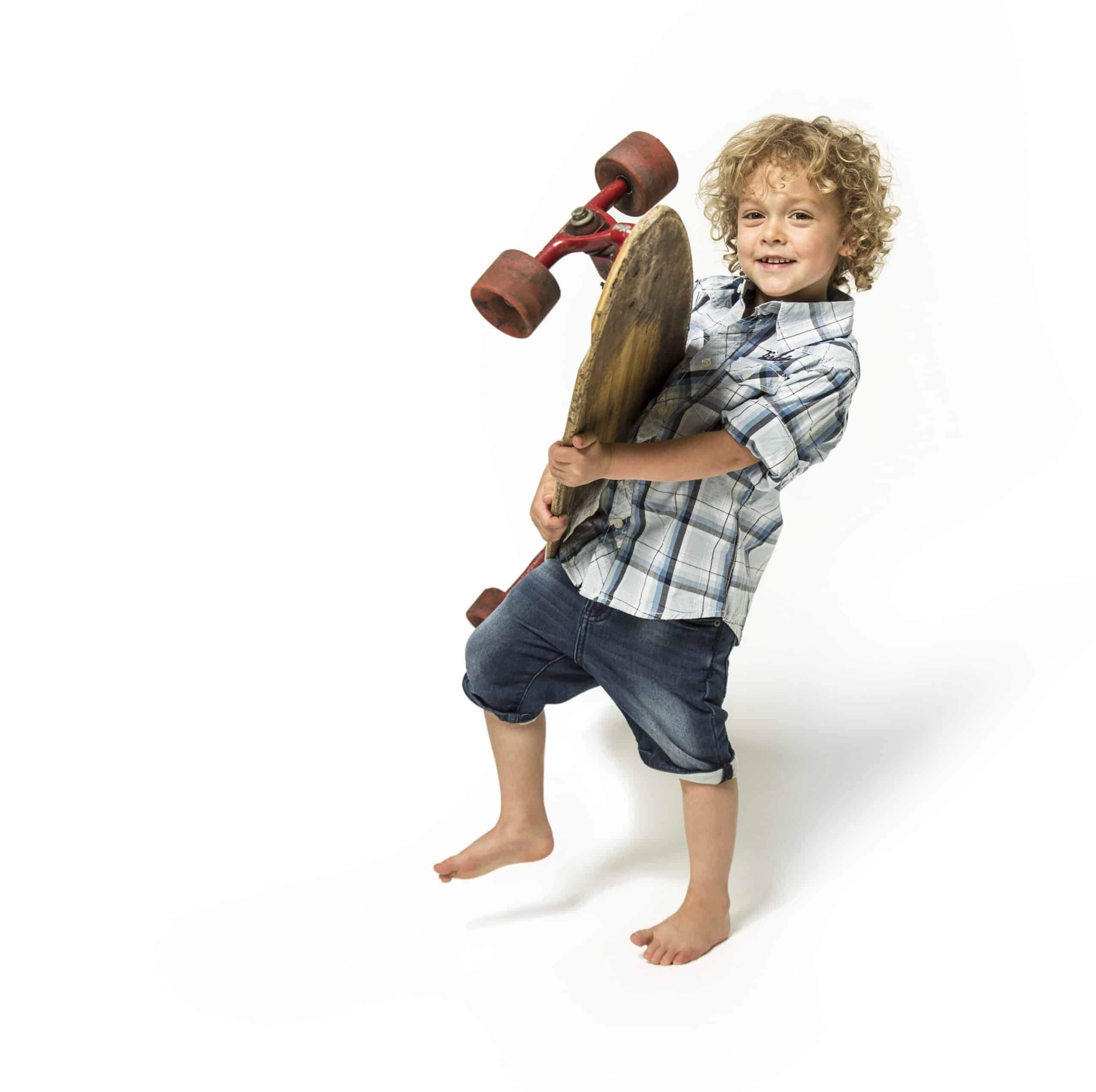 Ein kleines Kind mit lockigem Haar, das ein kariertes Hemd und hochgekrempelte Jeans trägt, steht barfuß und hält ein Skateboard. Das Kind hat einen fröhlichen Gesichtsausdruck und steht vor einem weißen Hintergrund. © Fotografie Tomas Rodriguez