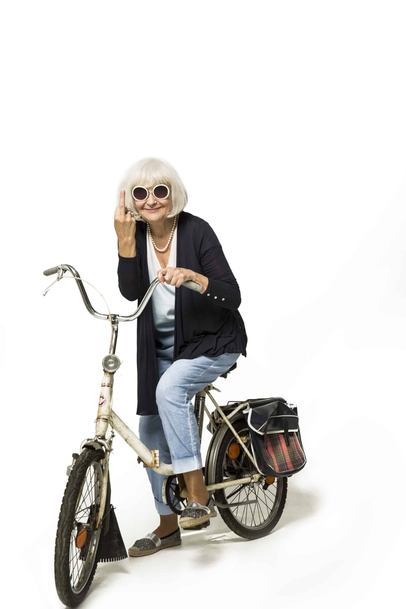 Eine ältere Frau mit weißem Haar, die eine schwarze Strickjacke, eine Sonnenbrille und hellblaue Jeans trägt, sitzt auf einem Vintage-Fahrrad mit einem Fahrradkorb hinten. Sie gestikuliert mit dem erhobenen Mittelfinger und strahlt damit eine verspielte oder rebellische Haltung aus. Der Hintergrund ist weiß. © Fotografie Tomas Rodriguez