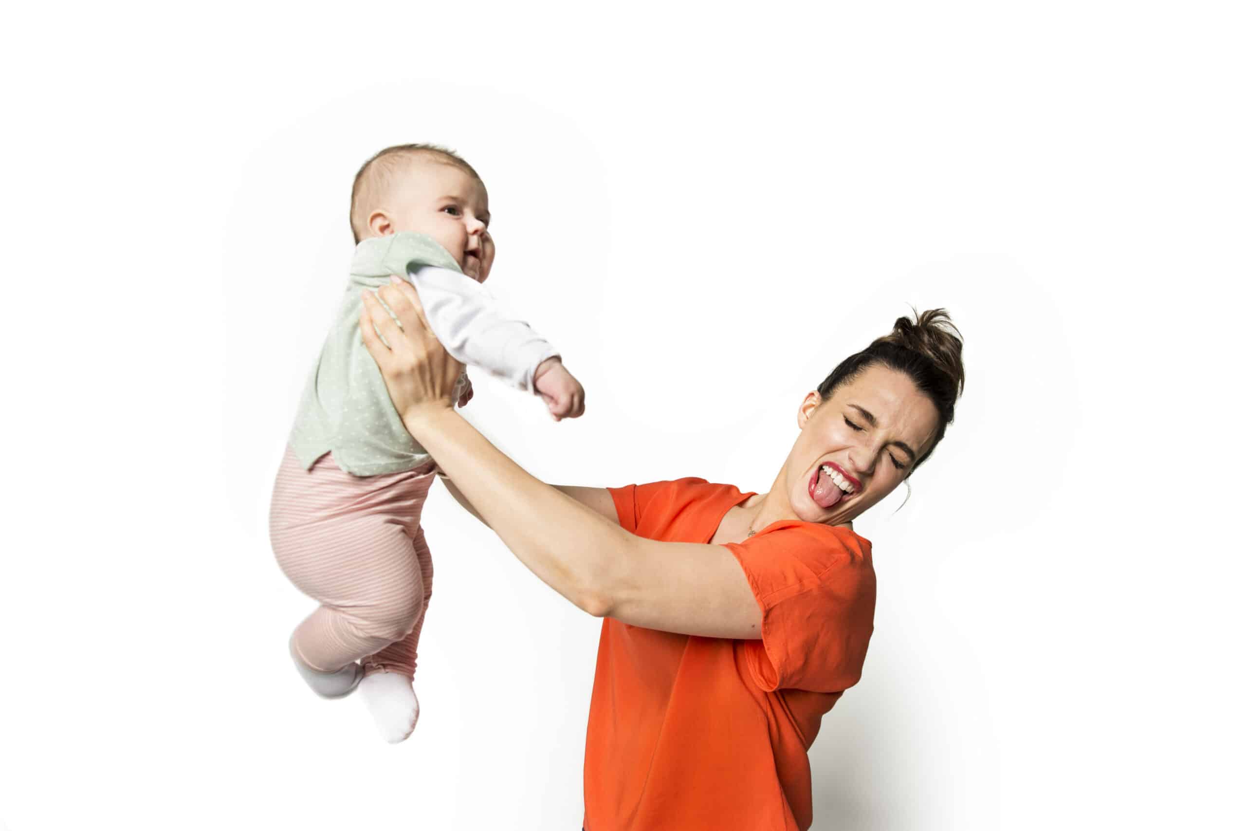 Eine Frau in einem orangefarbenen Hemd hält verspielt ein Baby, das ein grün-weißes Oberteil und eine rosa gestreifte Hose trägt. Die Frau schneidet ein lustiges Gesicht mit heraushängender Zunge und das Baby scheint zu kichern. Der Hintergrund ist schlicht weiß. © Fotografie Tomas Rodriguez